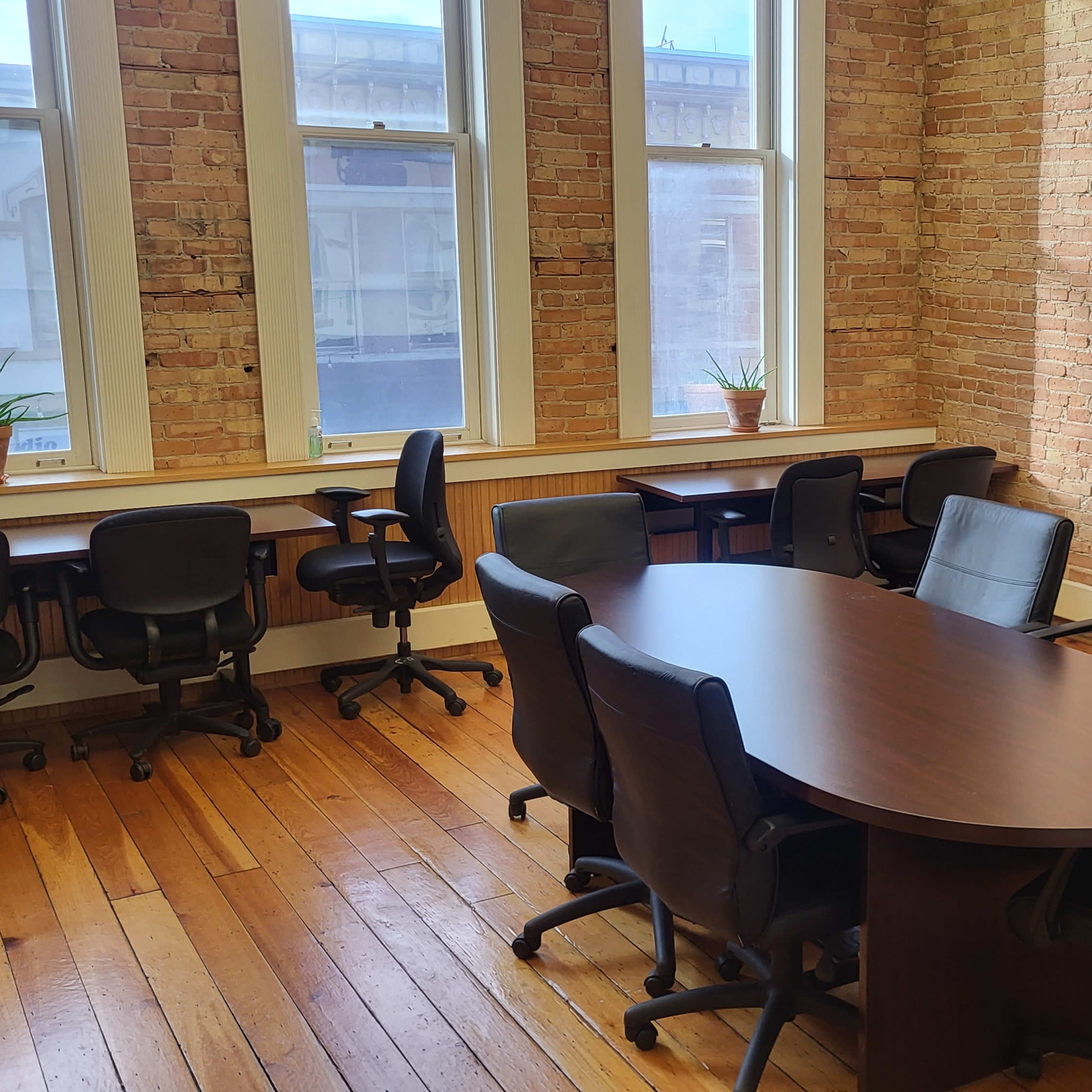 A conference room with a large oval table surrounded by eight black chairs, two potted plants on the windowsill, and a whiteboard on a stand against a brick wall.