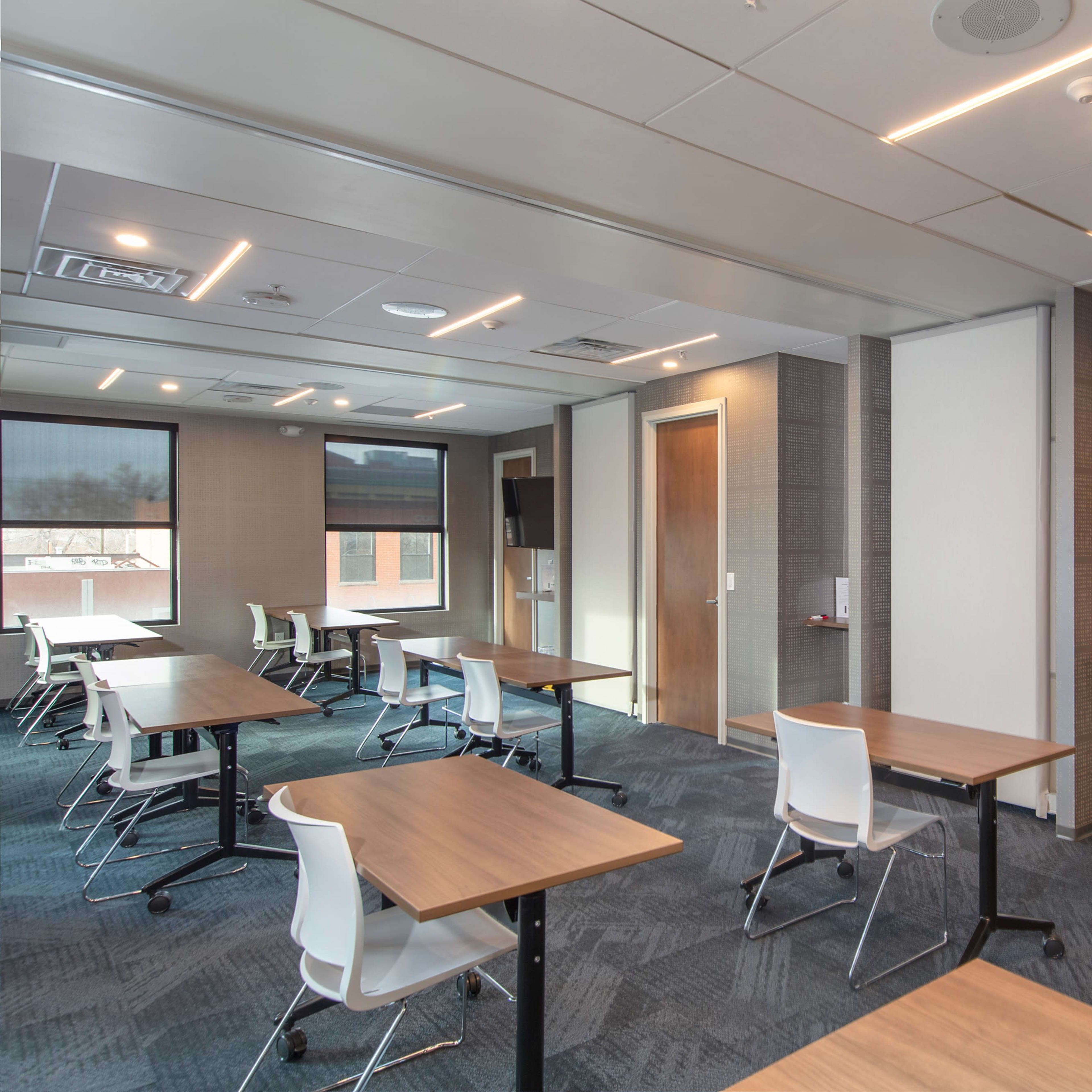 A modern classroom with several tables and chairs arranged neatly, along with a wall-mounted television and windows allowing natural light.