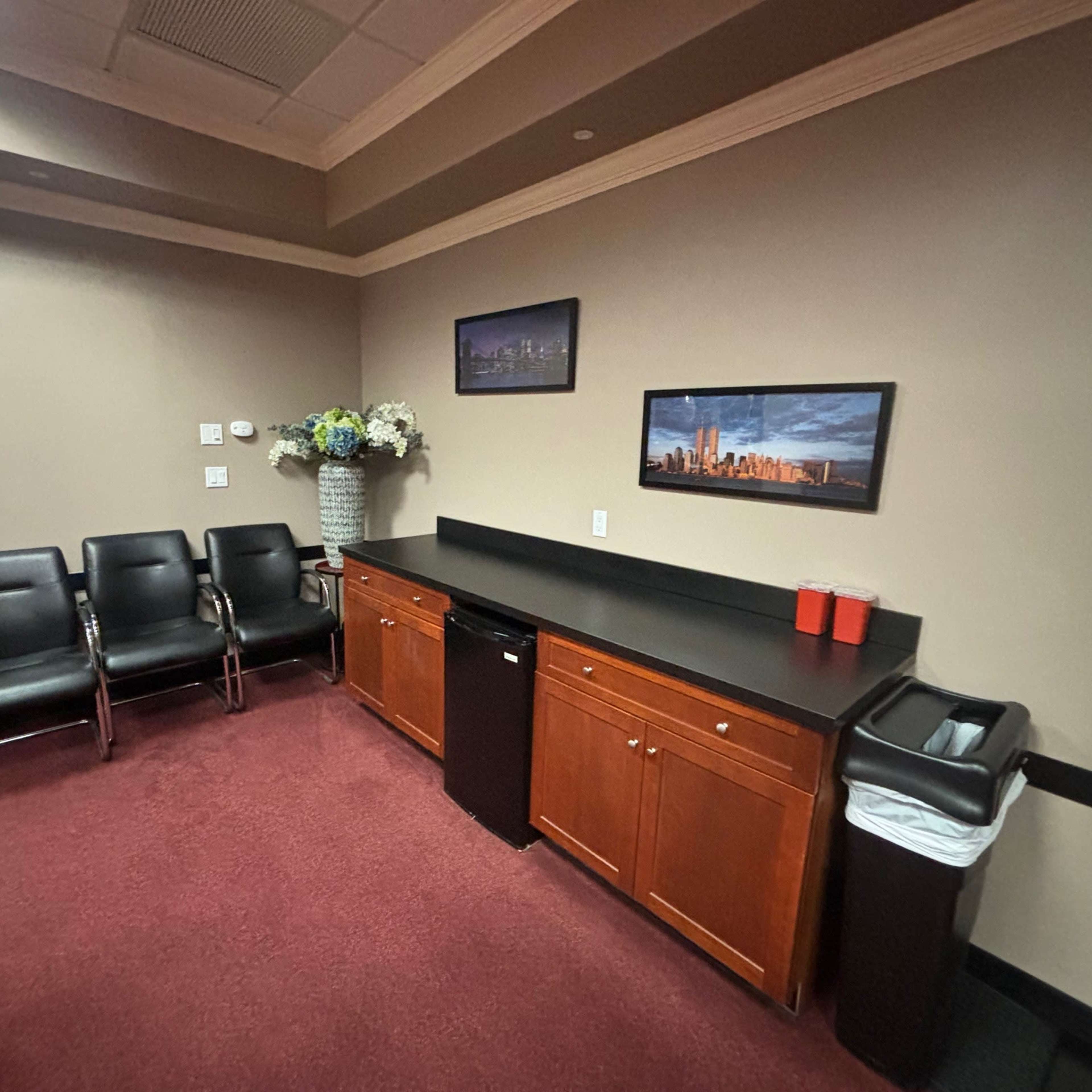 A small office waiting area with three black chairs, a countertop with two red cups, a small fridge, and a waste bin against a beige wall.