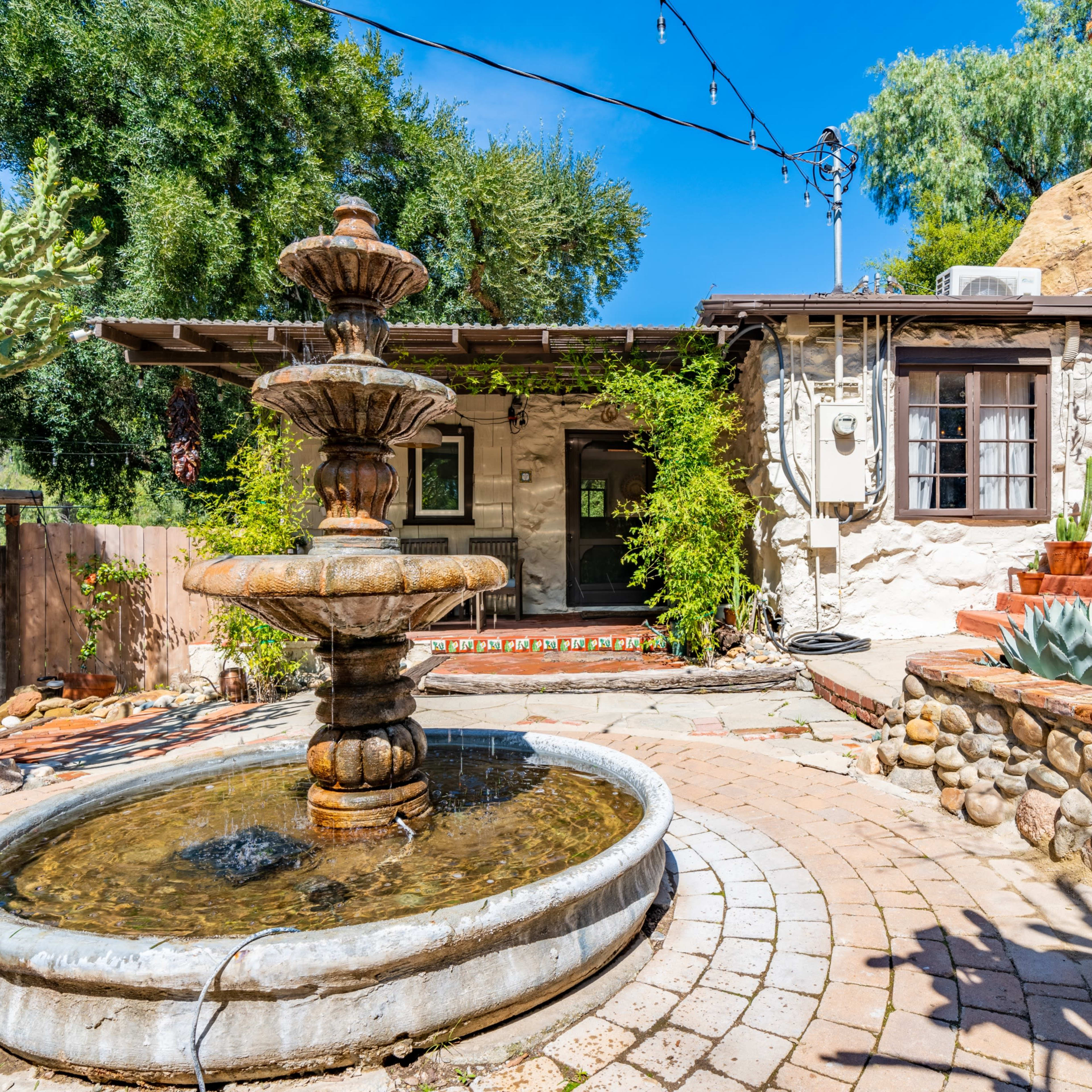 A stone fountain sits in a courtyard surrounded by desert plants and a rustic house featuring large windows and a staircase.