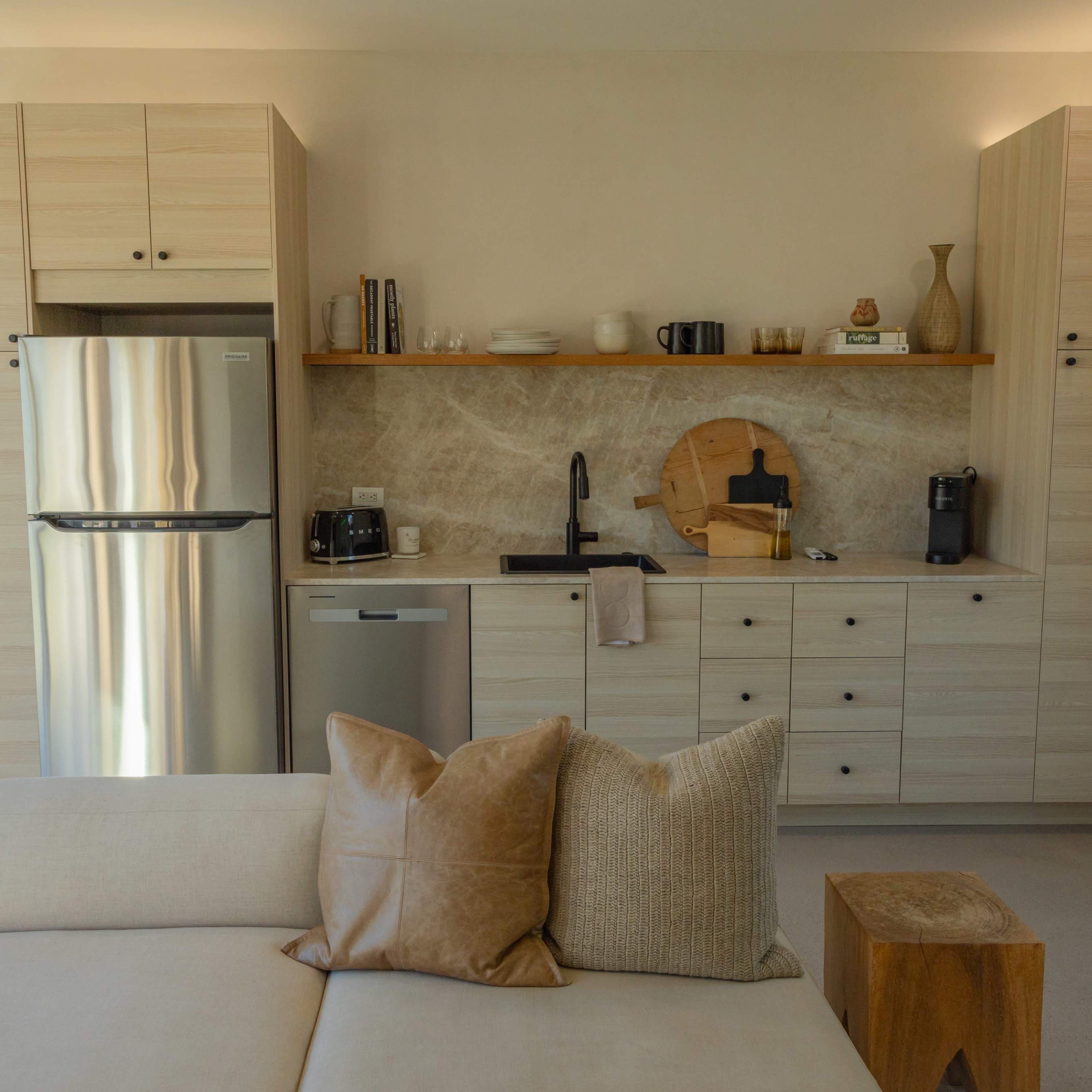 A modern kitchen features light wood cabinetry, a stainless steel refrigerator, a sink, and a small dining area with a sofa in the foreground.