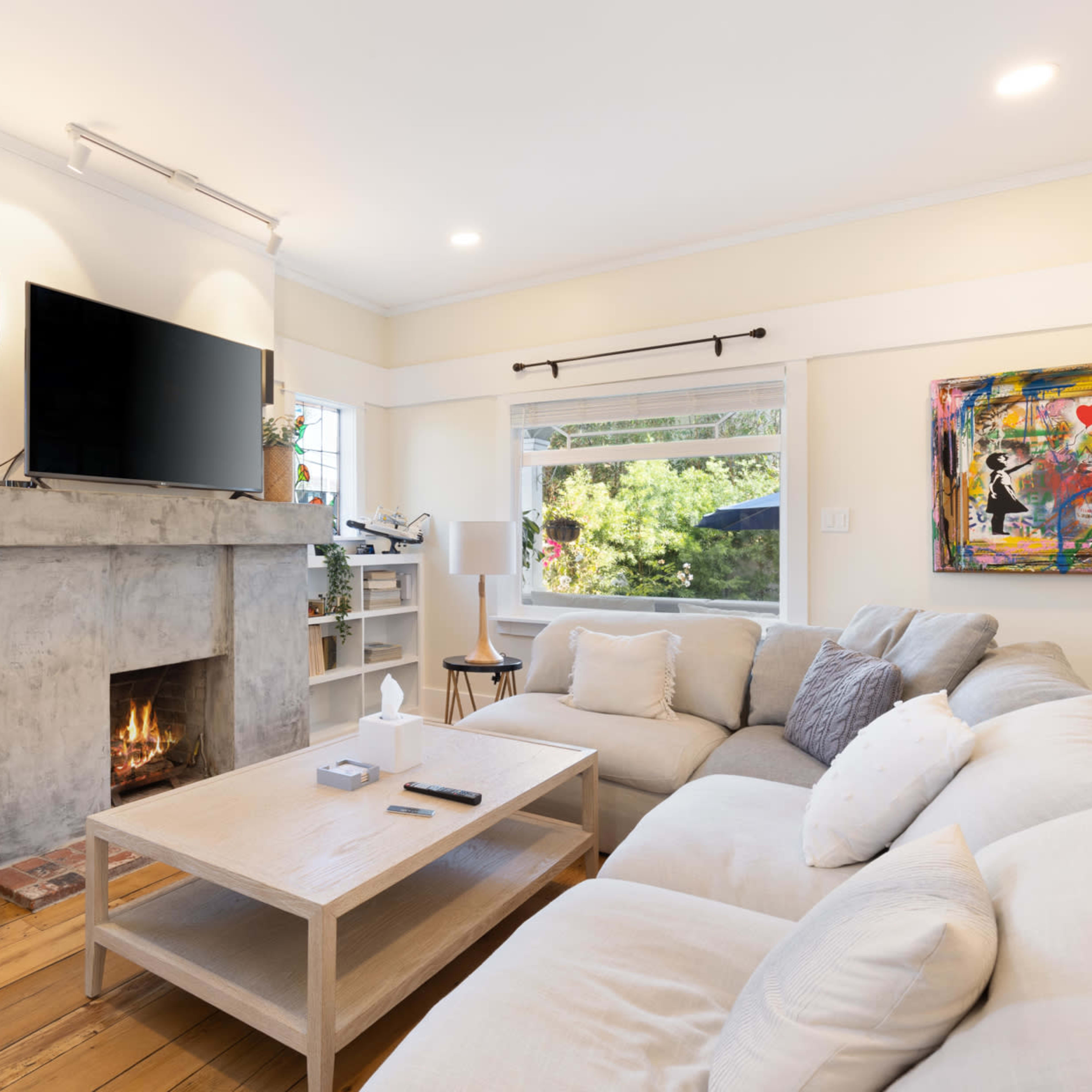 A cozy living room features a light-colored sectional sofa, a minimalist wooden coffee table, and a modern fireplace below a mounted TV.