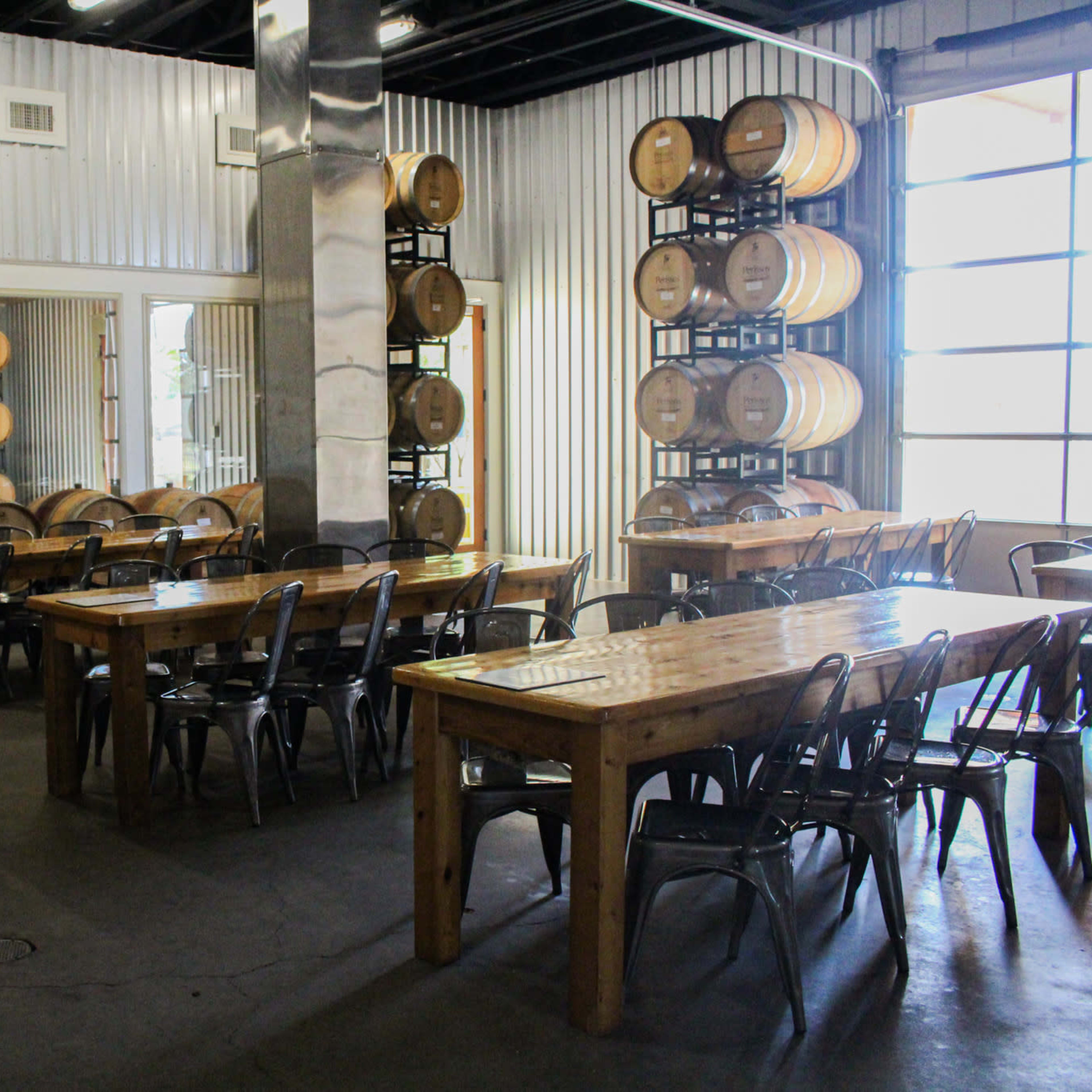 A spacious winery tasting room with wooden tables, metal chairs, and wine barrels stacked in the corner.