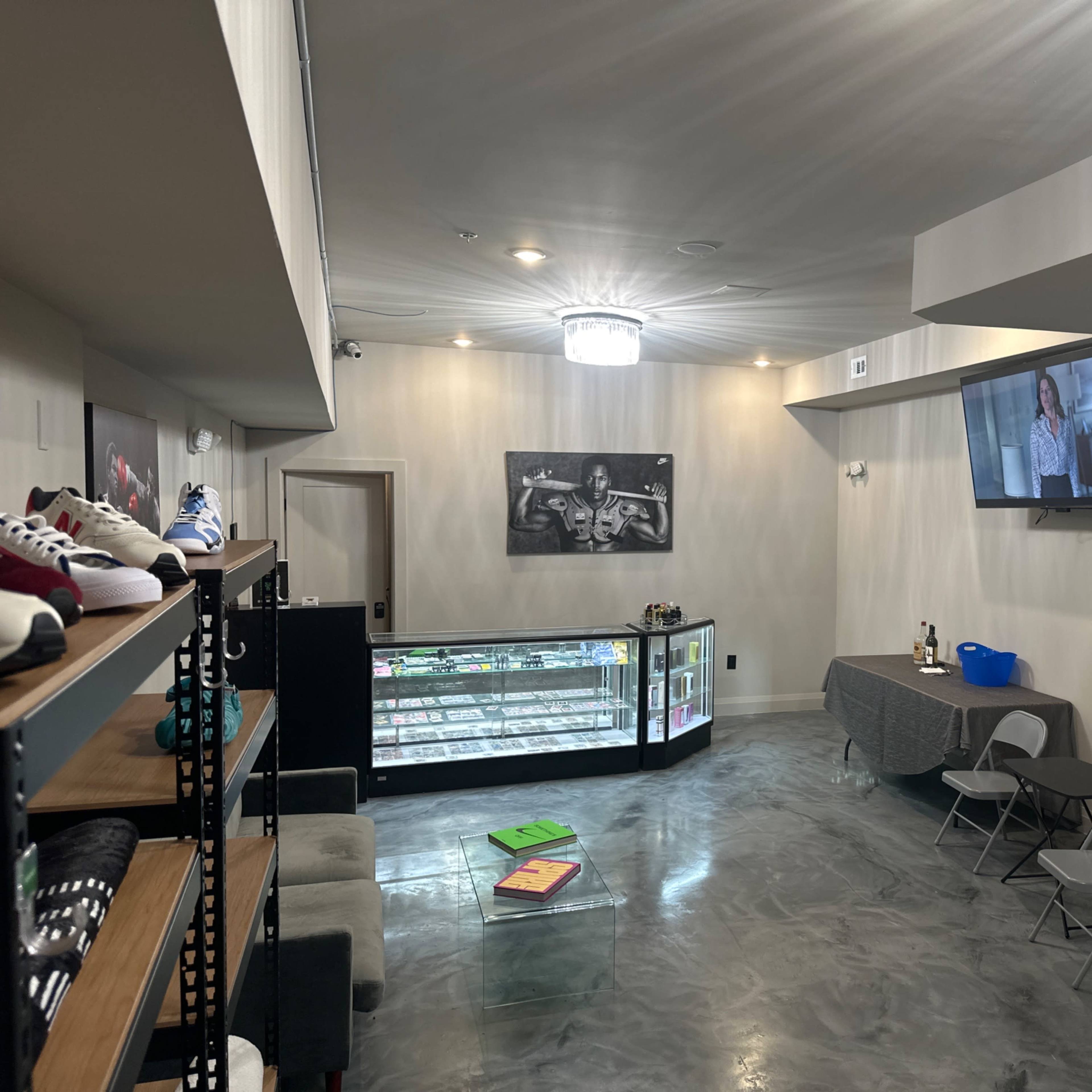 A modern retail space with concrete flooring, a display rack of shoes on the left, a table with chairs in the center, and a wall-mounted television in the corner.