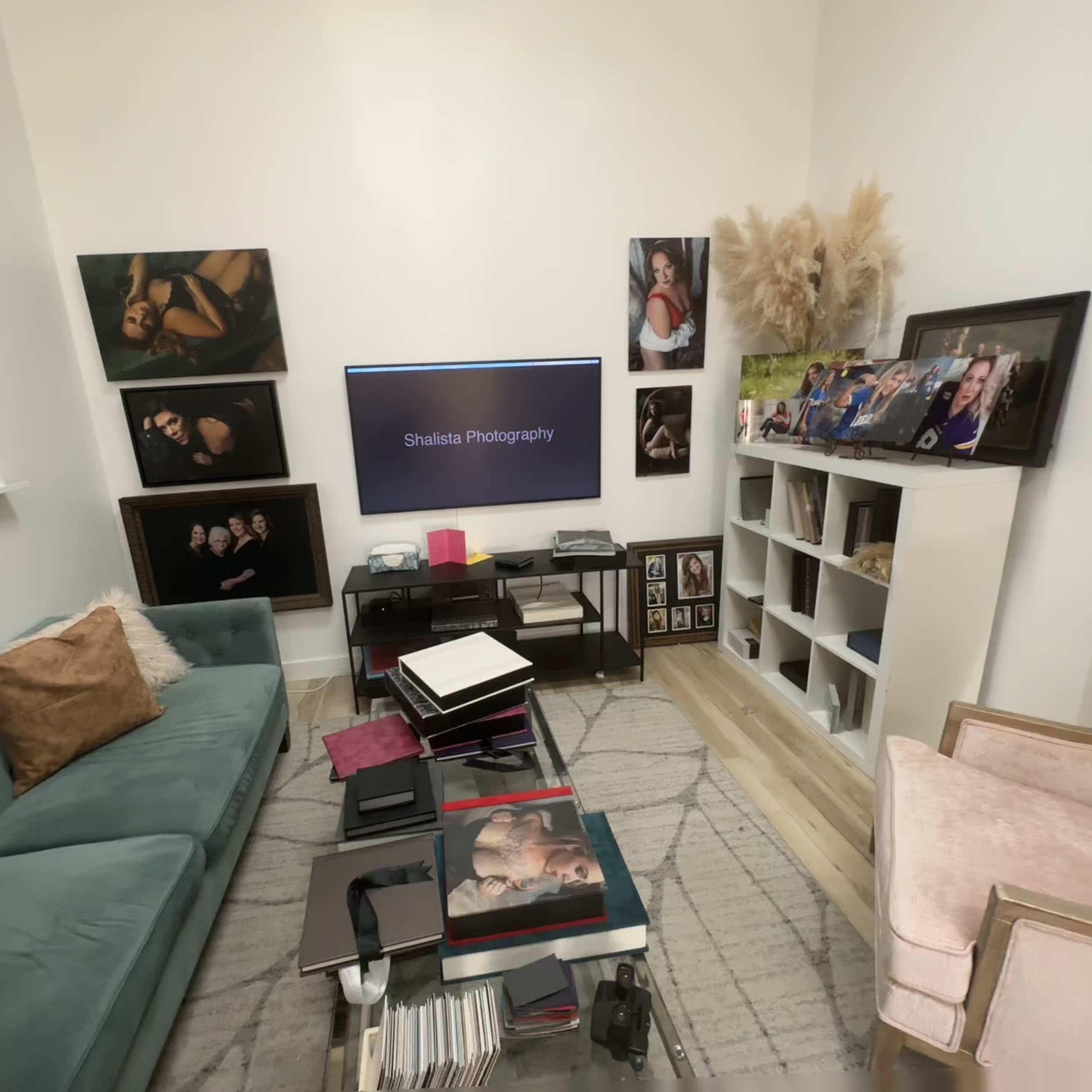 A cozy photography studio with a green couch, a television displaying "Shakira Photography," various framed photographs on the walls, and a bookshelf filled with photo albums.