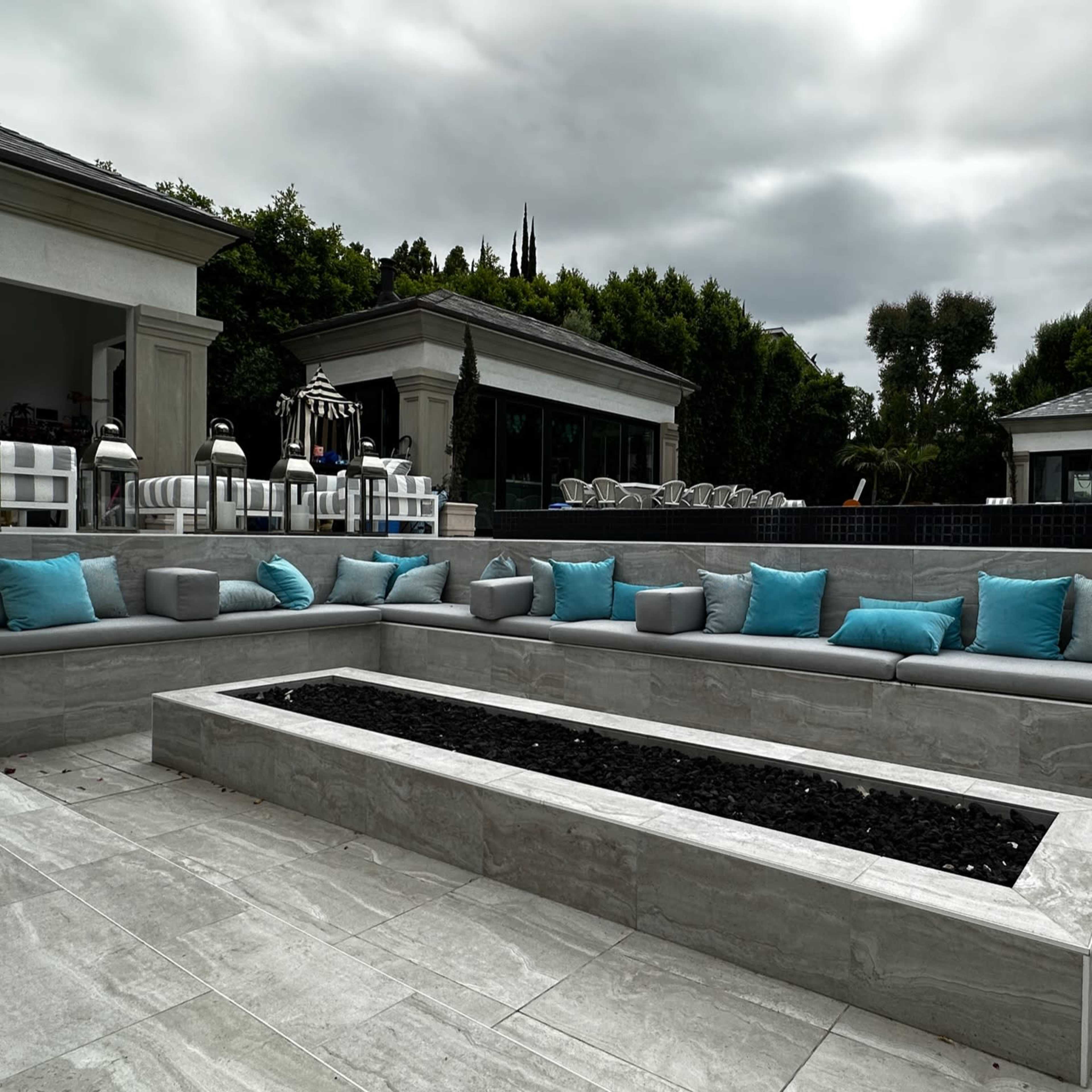 A modern outdoor seating area features gray stone benches with turquoise accent pillows surrounding a low stone firepit.