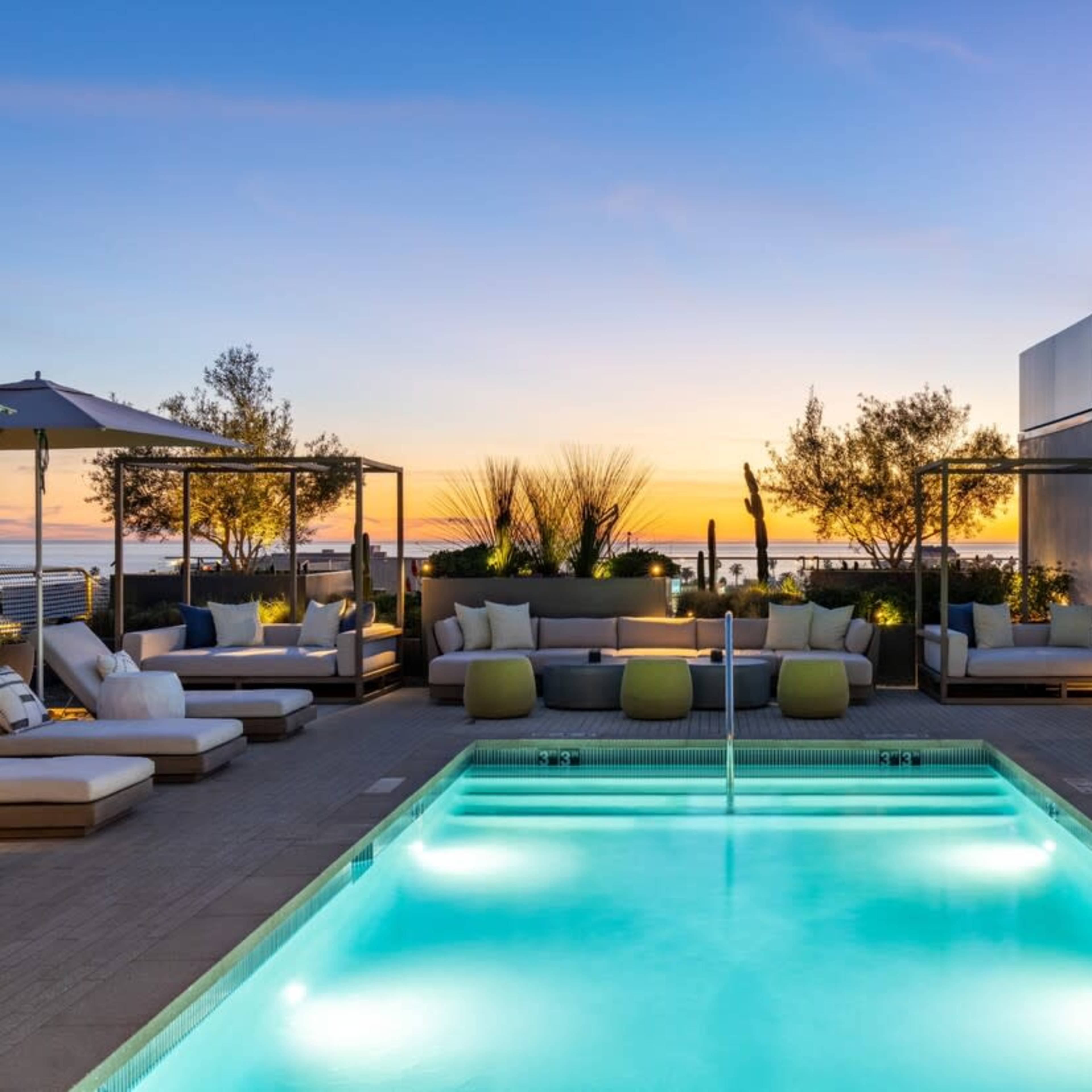 A modern rooftop terrace features a swimming pool, lounge seating, and umbrellas against a sunset backdrop.