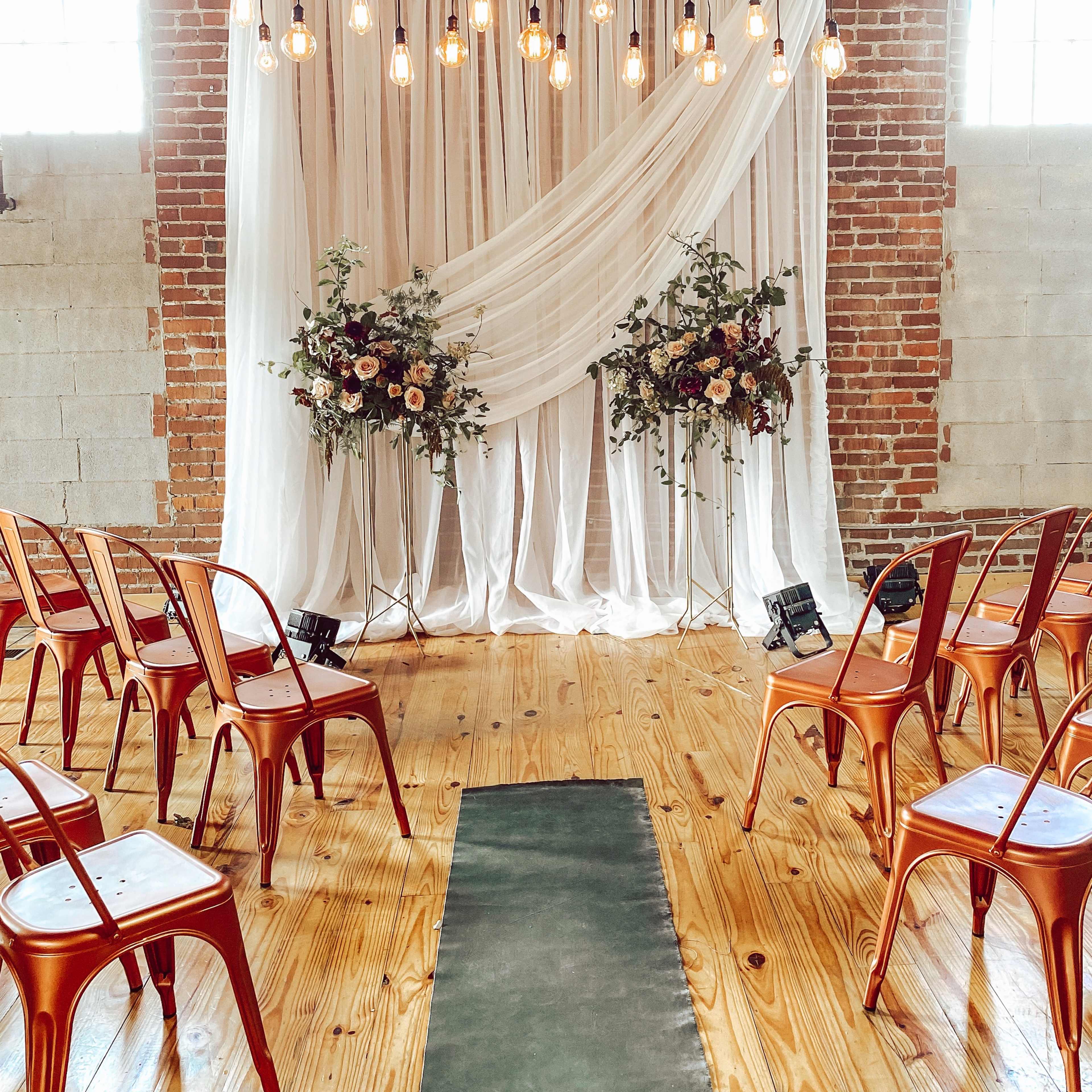 A wedding ceremony setup with a decorative fabric backdrop, hanging lights, floral arrangements, and rows of chairs arranged on a wooden floor.