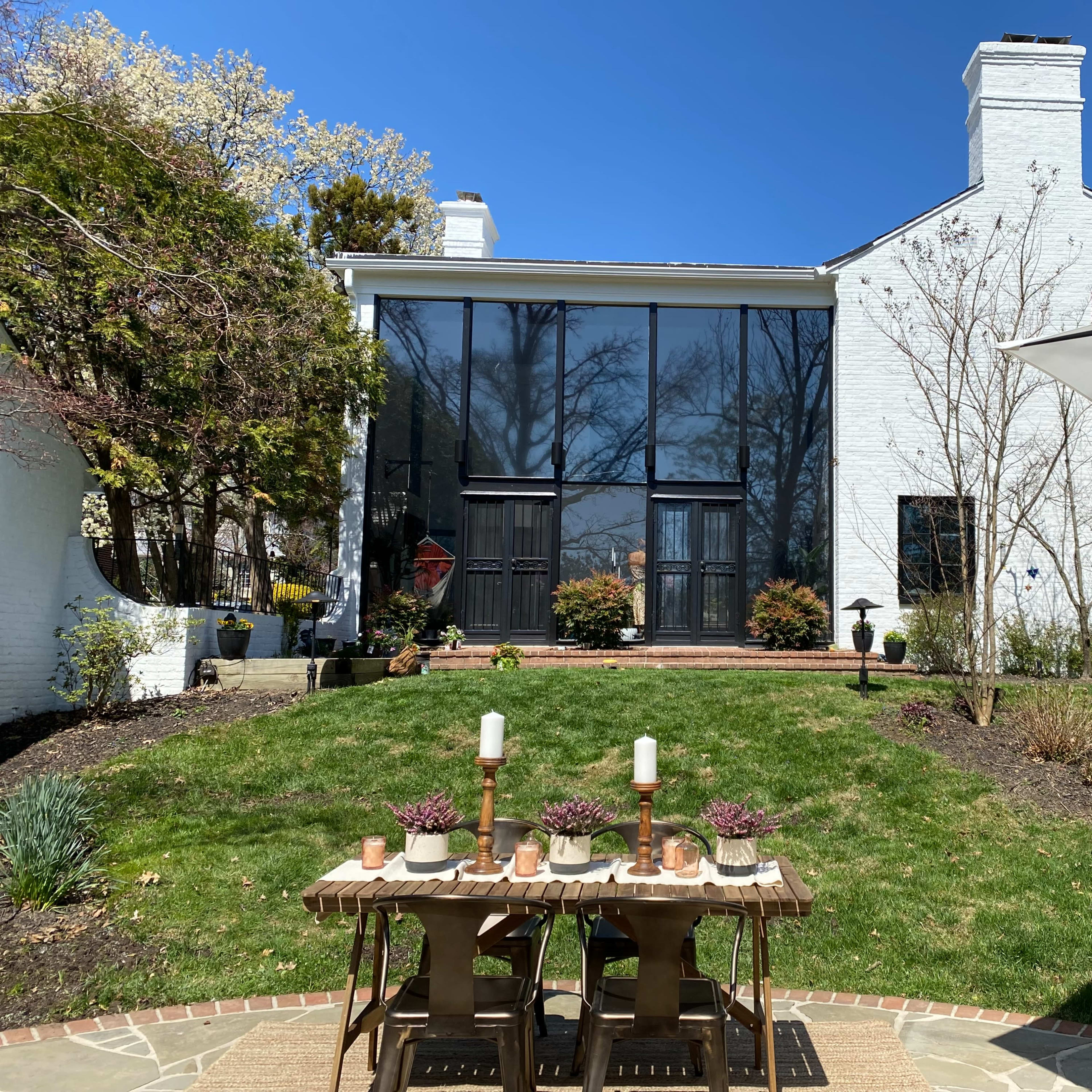 A modern white house with large glass doors is set against a clear blue sky and surrounded by greenery, with a wooden table placed on the lawn.