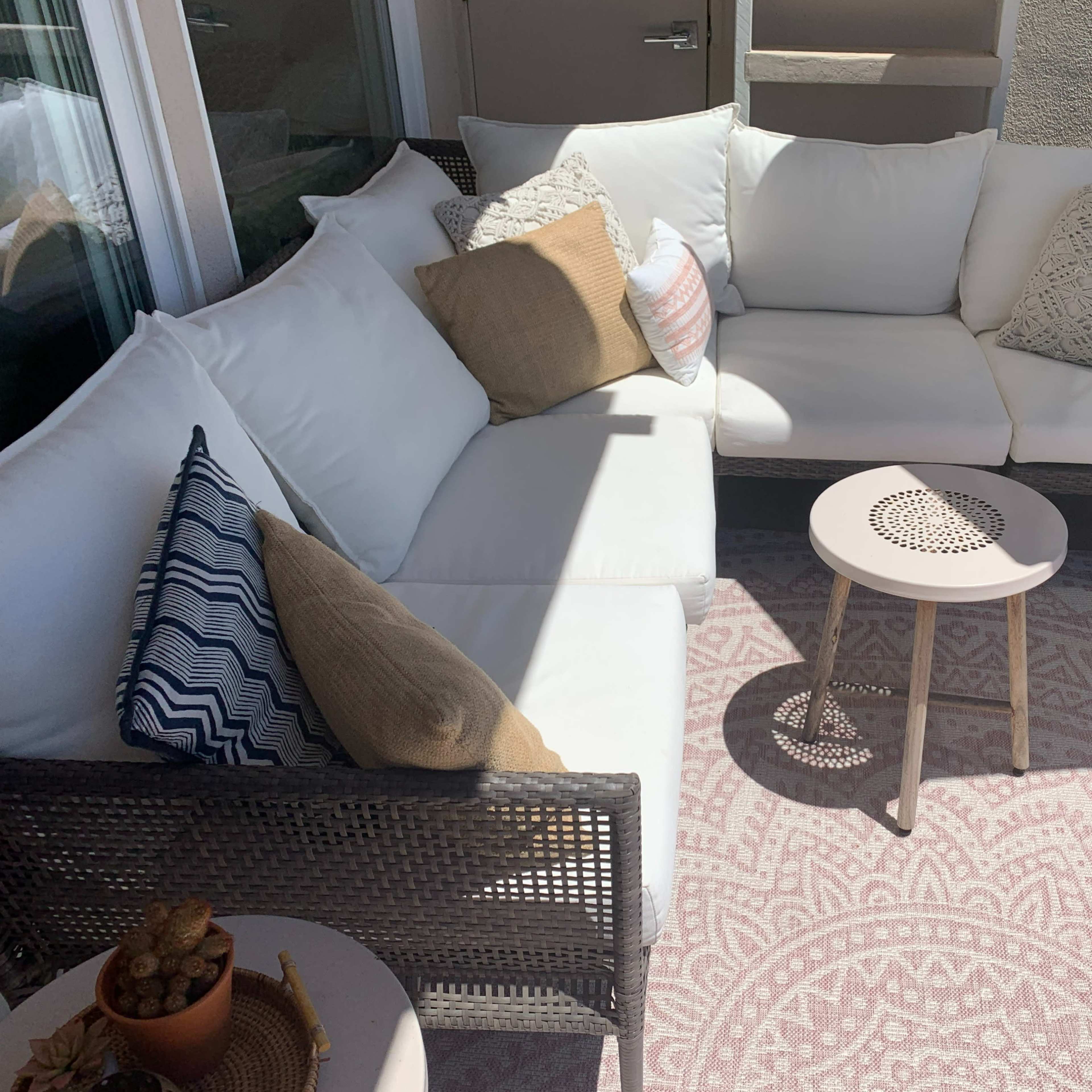 A cozy outdoor seating area featuring a sectional couch with white cushions, decorative pillows, and a small round table on a patterned rug.