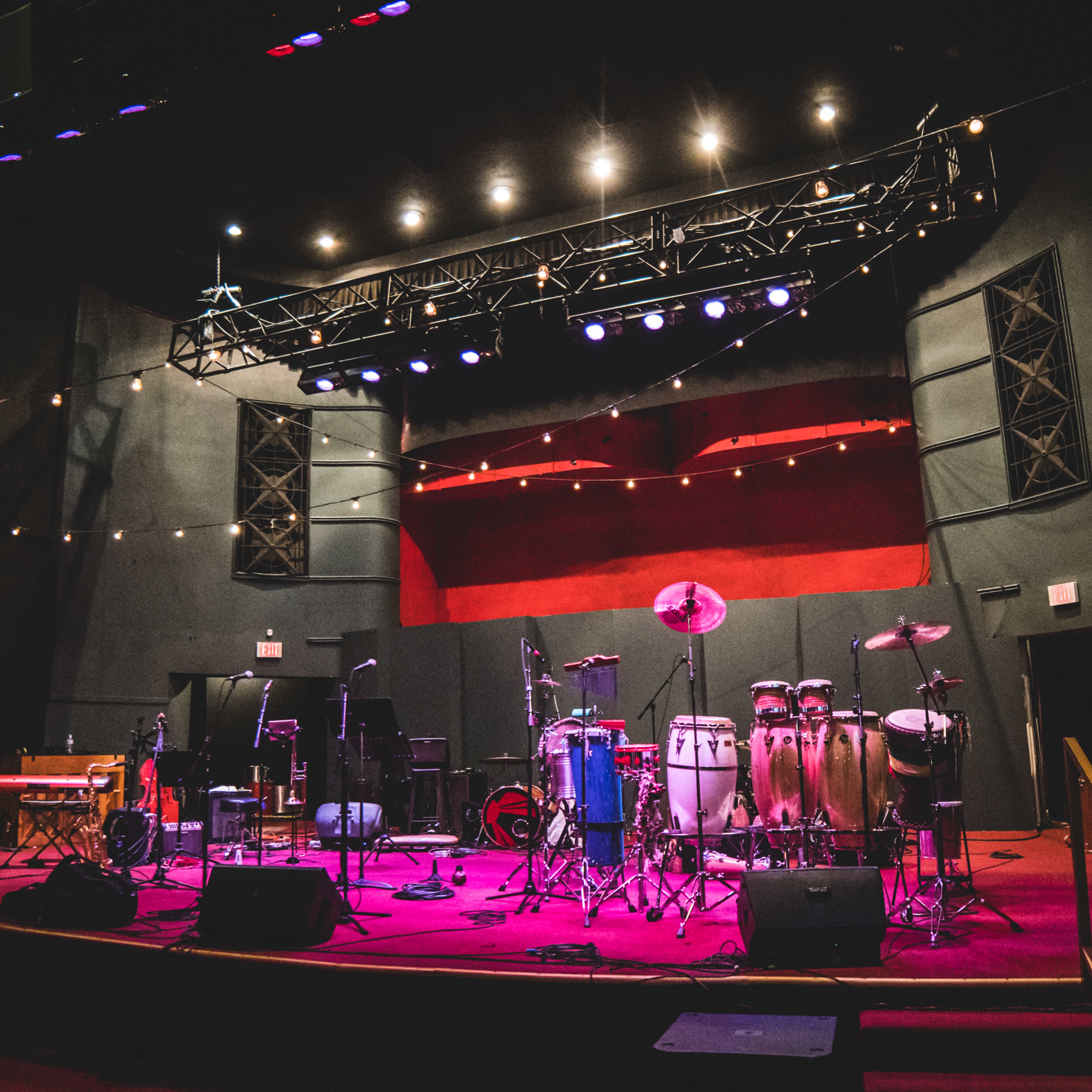 A stage set up with various musical instruments, including congas and a drum set, is illuminated by colorful stage lights.