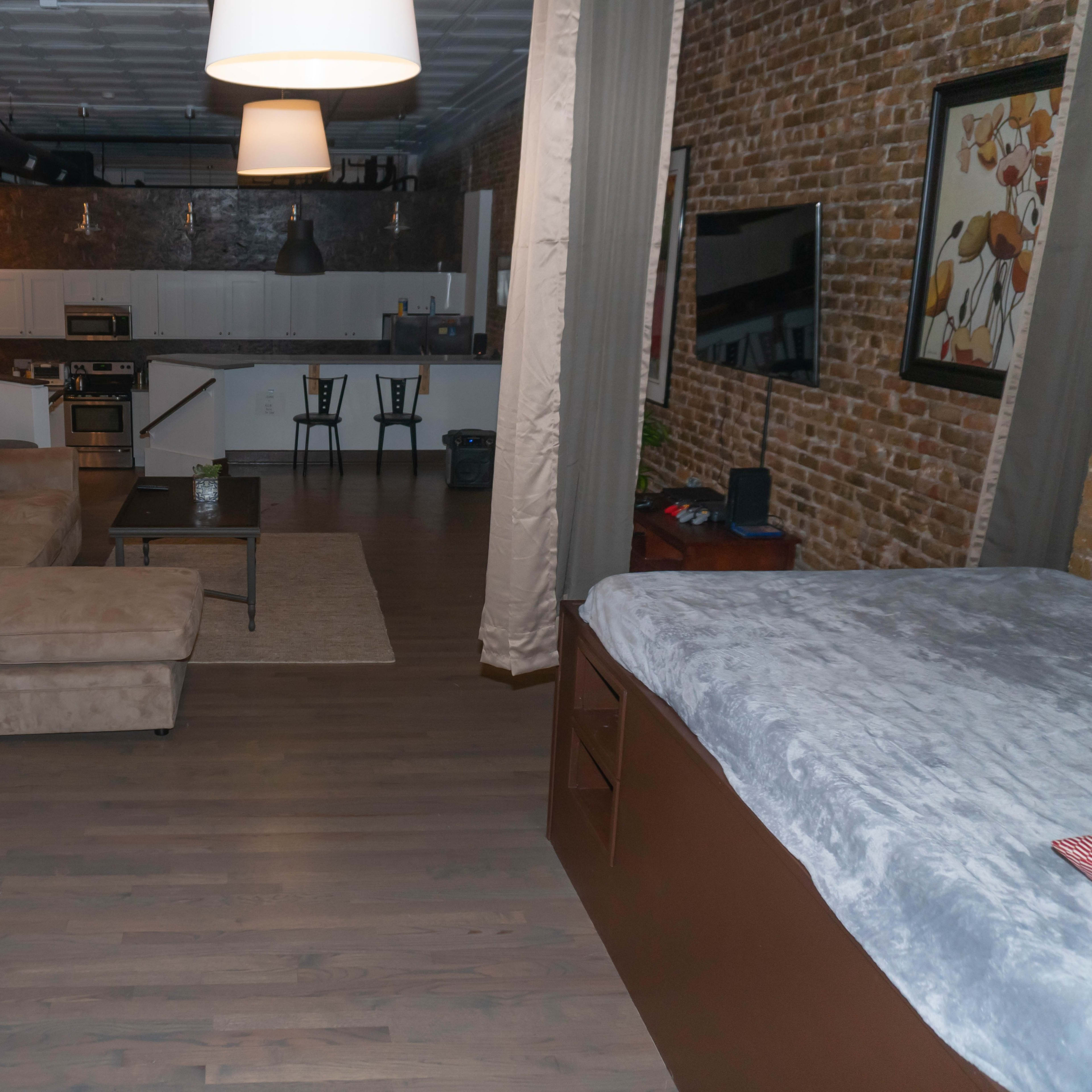 An open loft-style living space featuring a living area with a couch, a kitchenette in the background, and a bed area with a grey blanket.