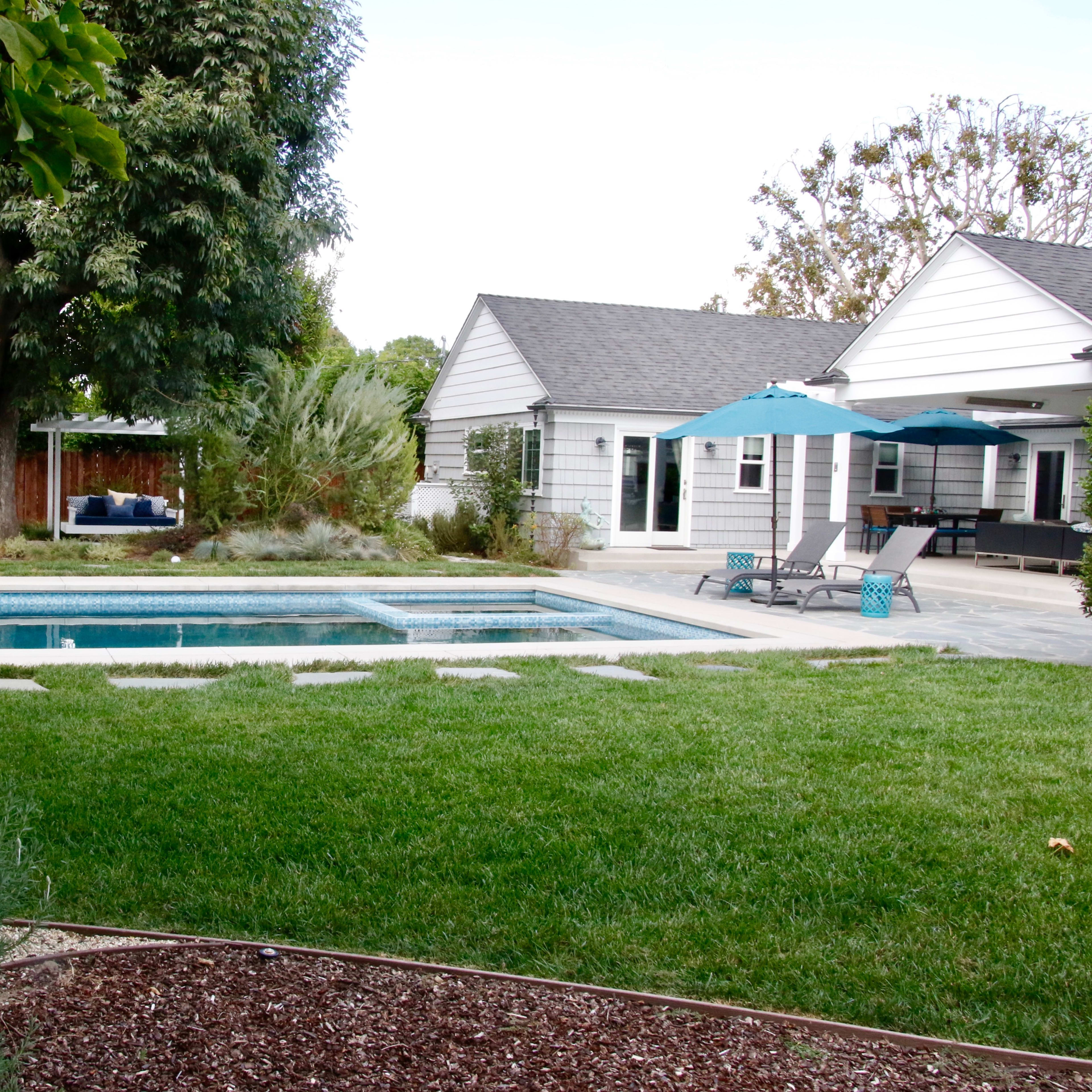 A neatly landscaped backyard features a swimming pool, bordered by grassy areas and stone pathways, with a patio and a nearby shaded seating area.