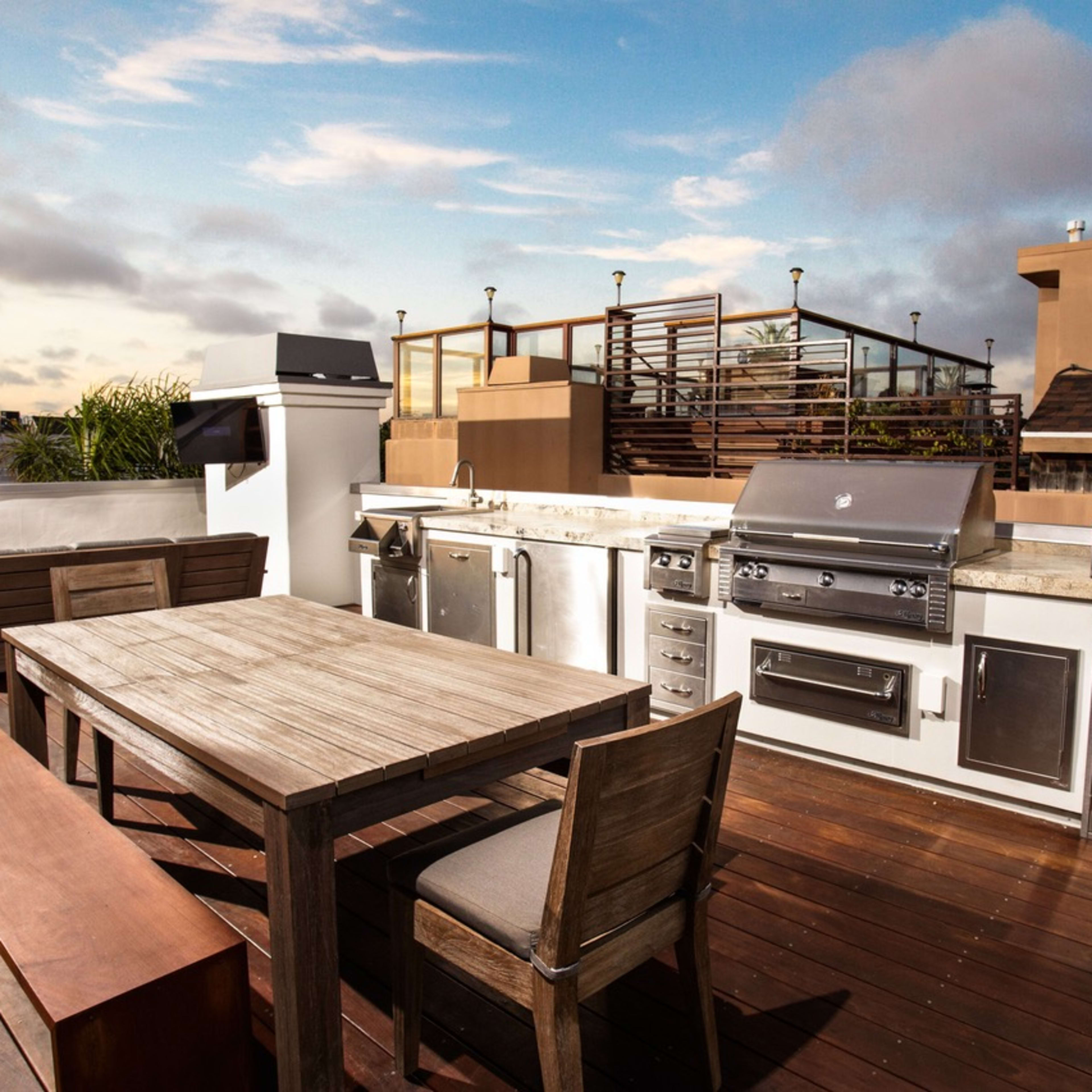 A rooftop patio with a barbecue grill, outdoor kitchen appliances, a wooden dining table, and seating areas surrounded by a skyline view.