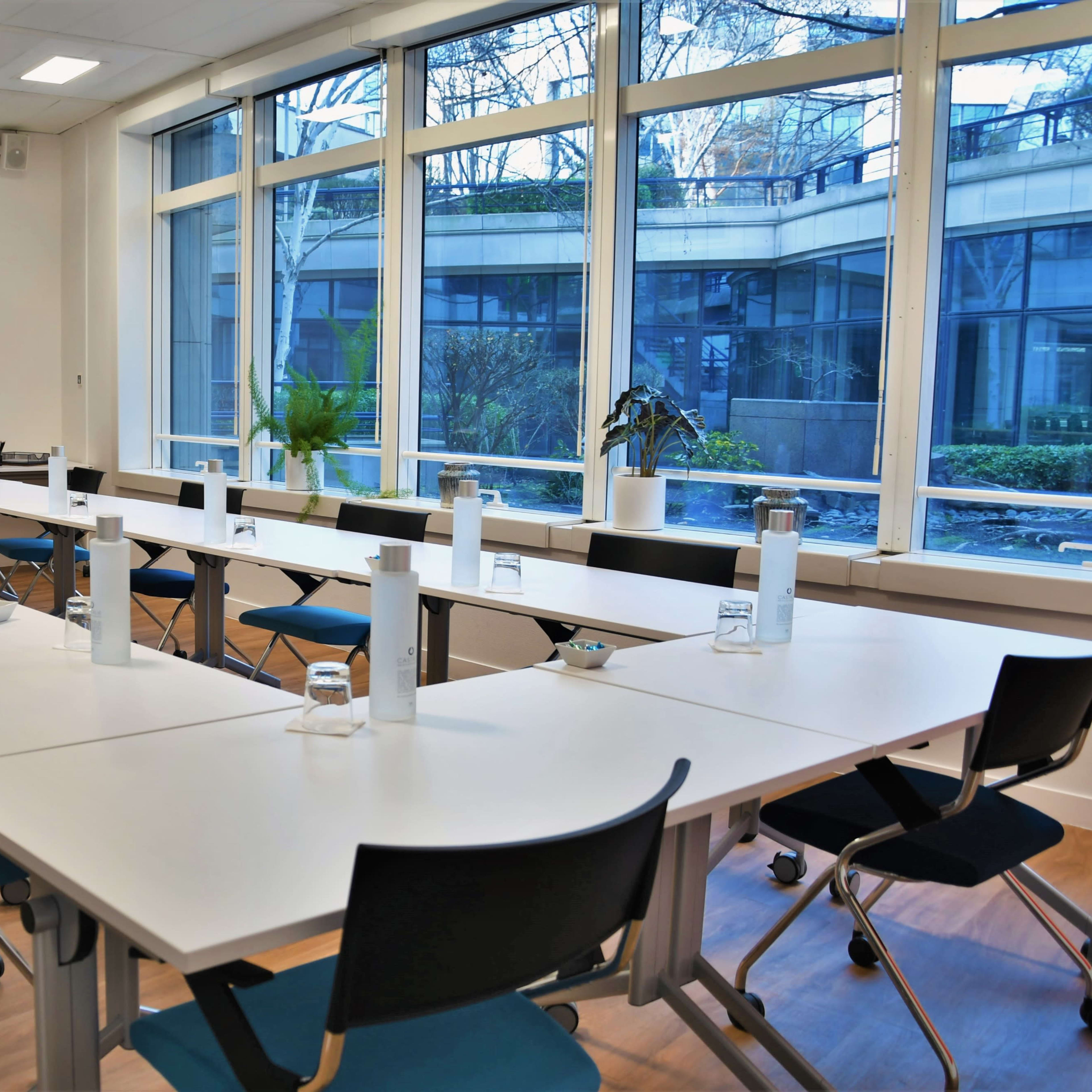 A large, modern conference room features a U-shaped arrangement of tables with chairs, glass windows, and plants in the background.