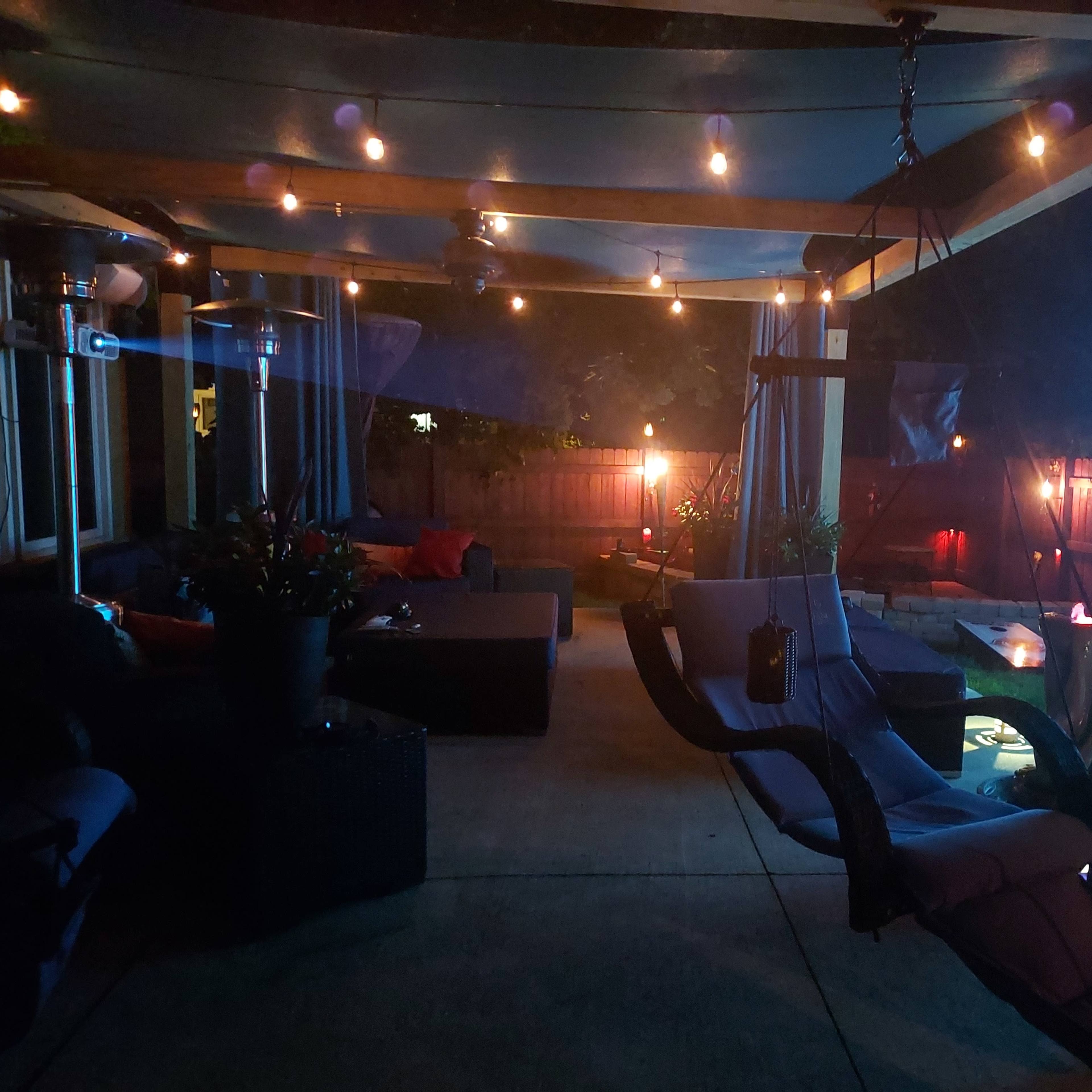 A cozy outdoor patio area with a lounging space, string lights, heating elements, and a projector screen displaying a blue image.