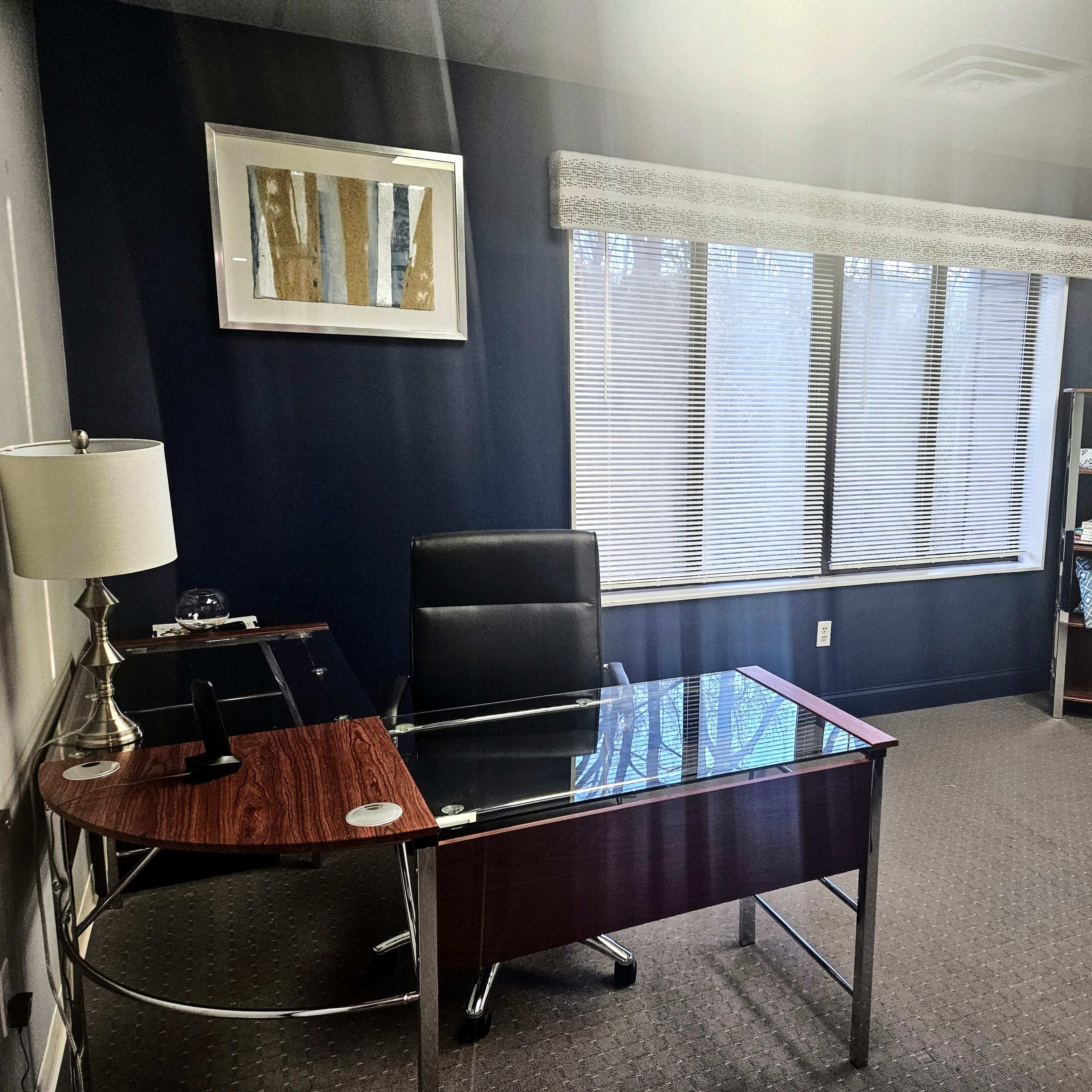 A minimalist office space featuring a glass-top desk, a black chair, a blue couch, and a window with blinds.