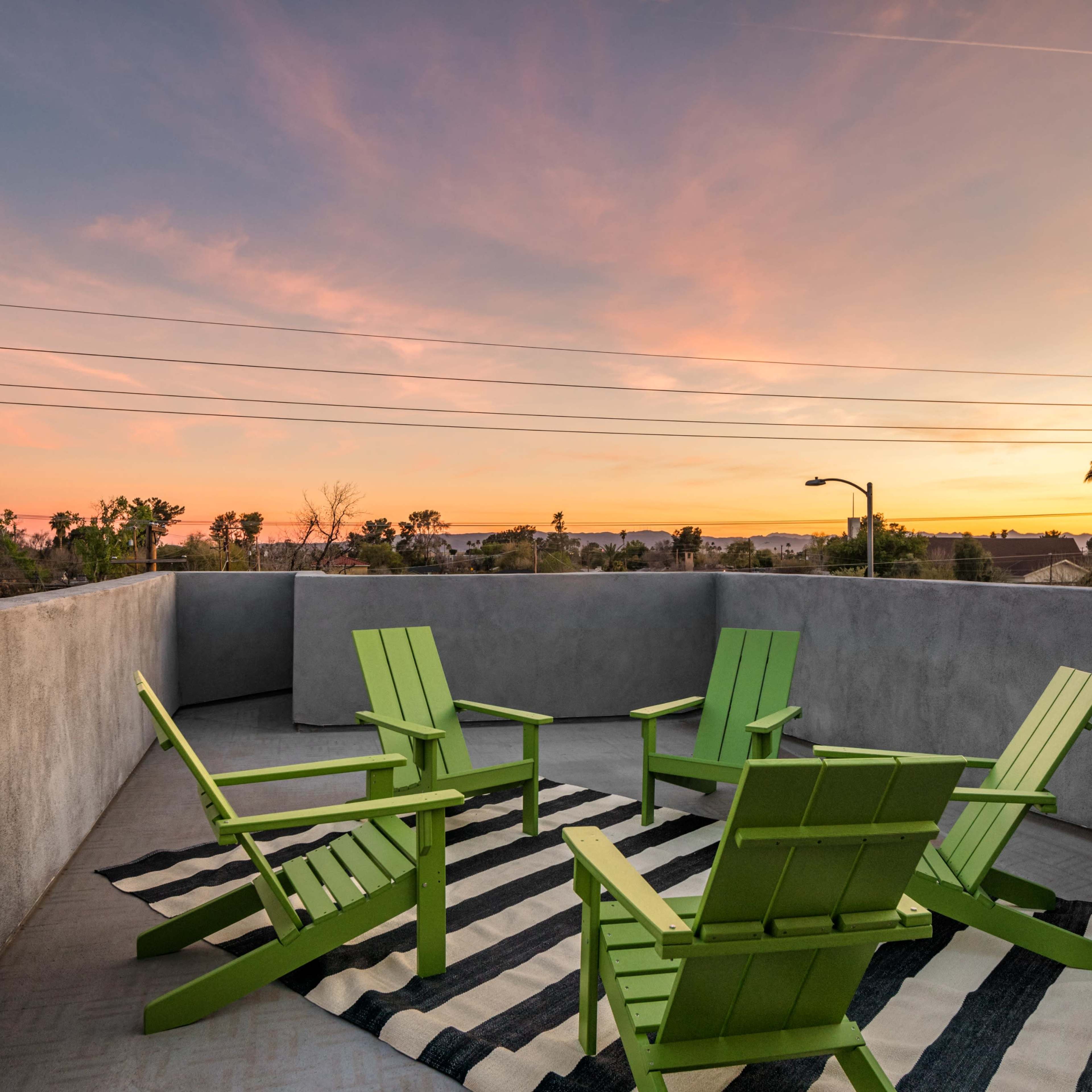 A rooftop patio features four green chairs arranged around a striped rug, with a colorful sunset in the background.