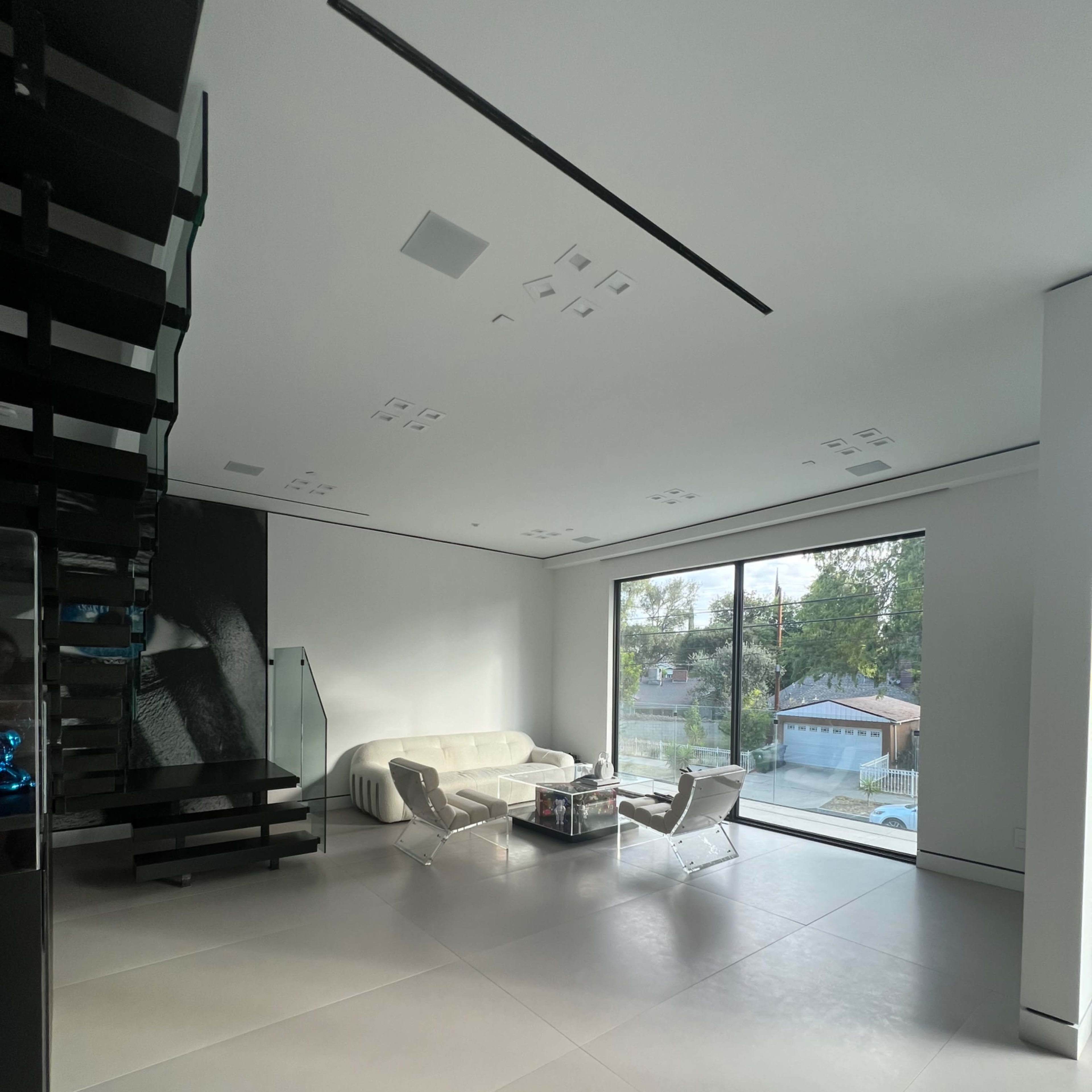 A modern living room with a minimalist design, featuring a white sofa, two sleek chairs, a coffee table, and large windows overlooking a garden.