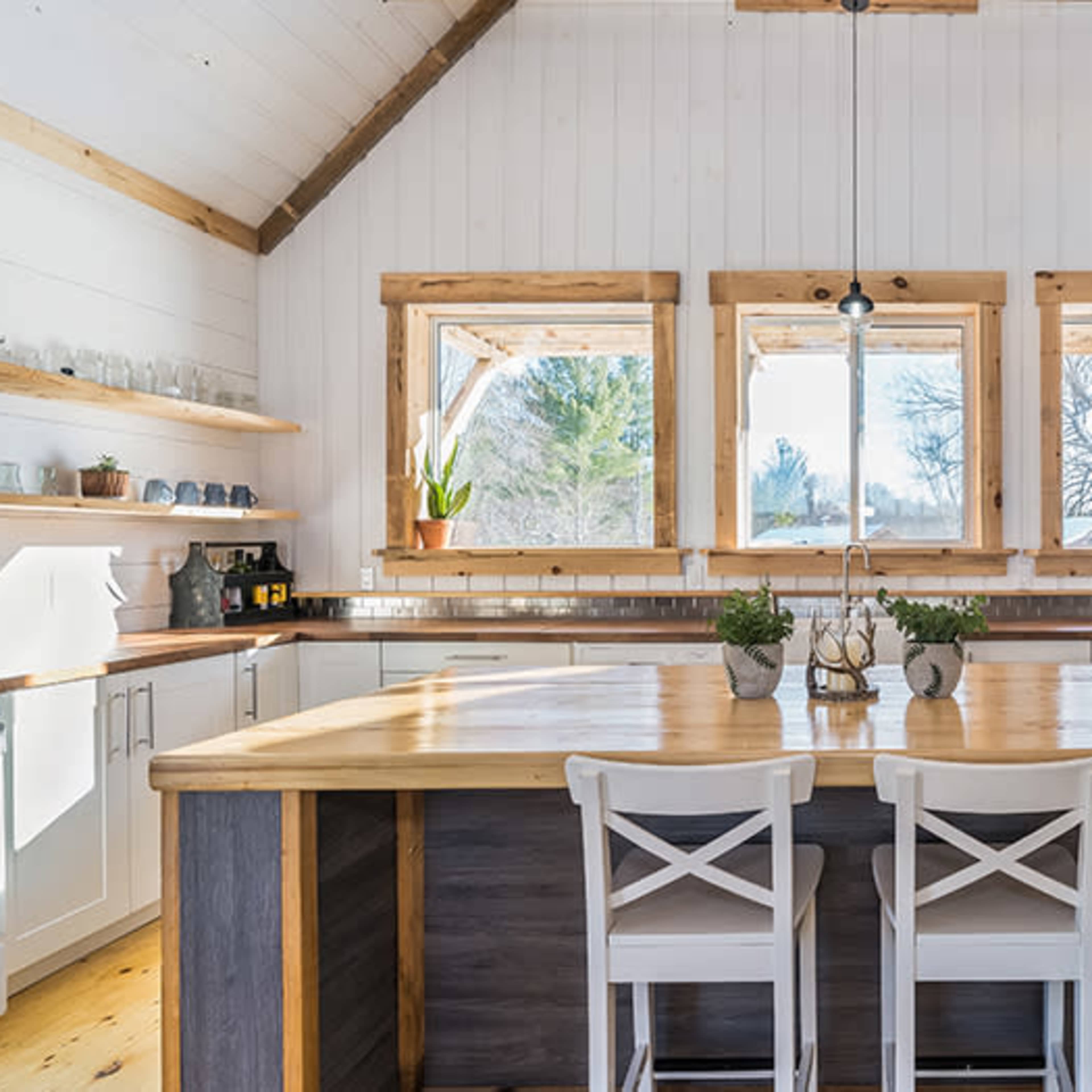 A modern kitchen features a central wooden island with two white chairs, surrounded by white cabinets and large windows that let in natural light.