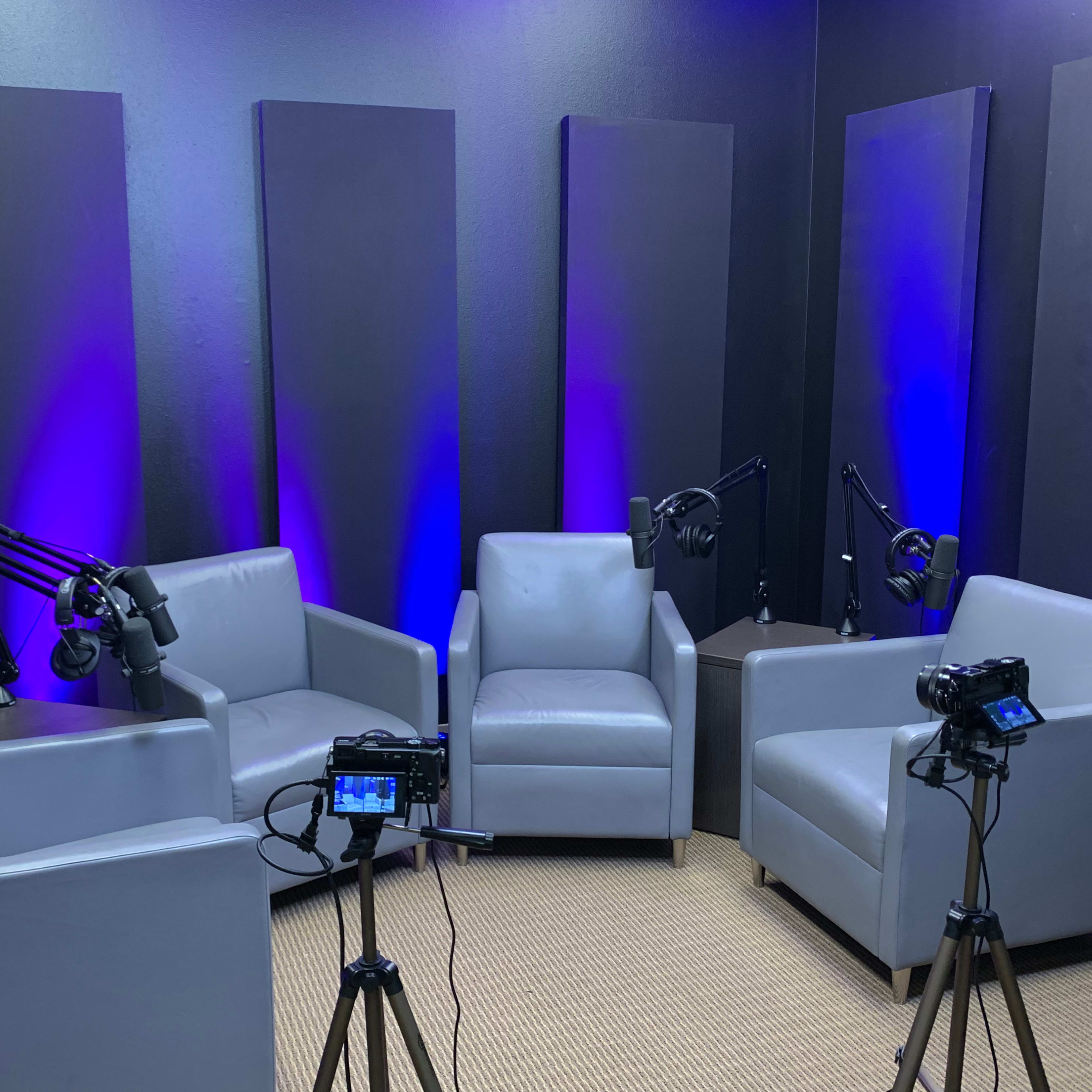 A podcast studio with four gray chairs, two microphones on adjustable arms, and two cameras set up on tripods.