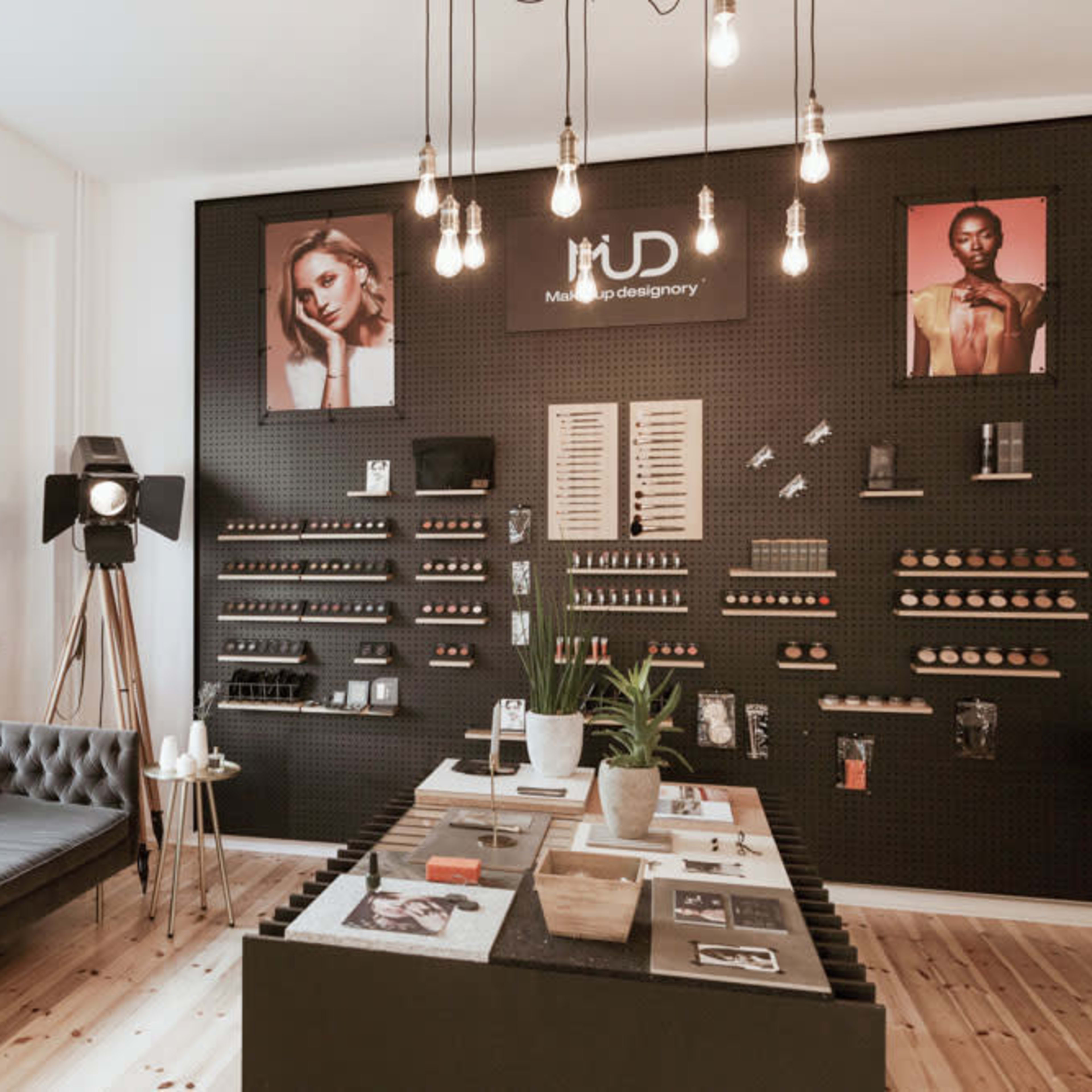 A modern beauty studio features a dark pegboard wall displaying makeup products, a central table with various items, and a cozy seating area.