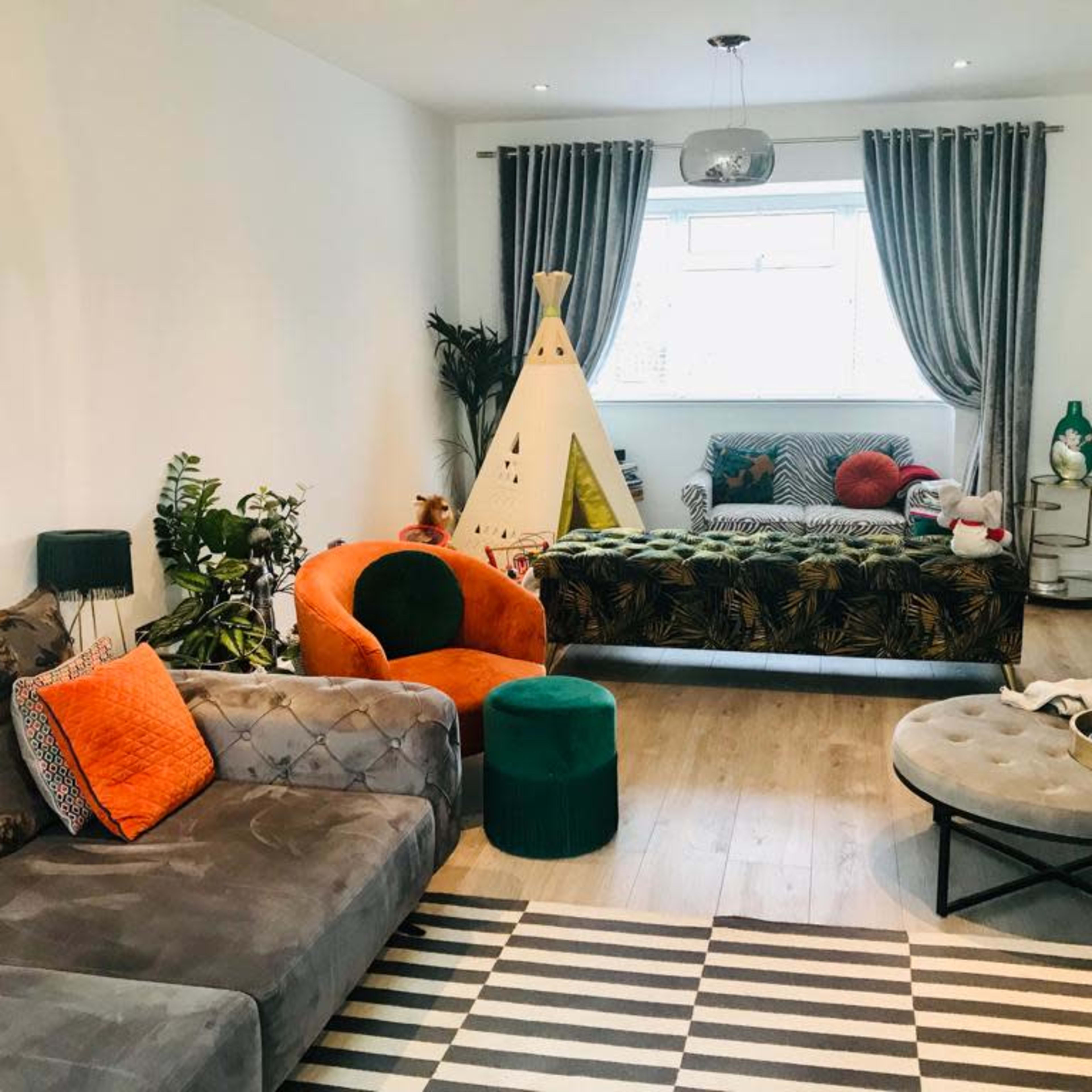 The living room features a mix of seating options, including a gray sofa, an orange chair, and a patterned couch, with a children's teepee and various plants accentuating the space.