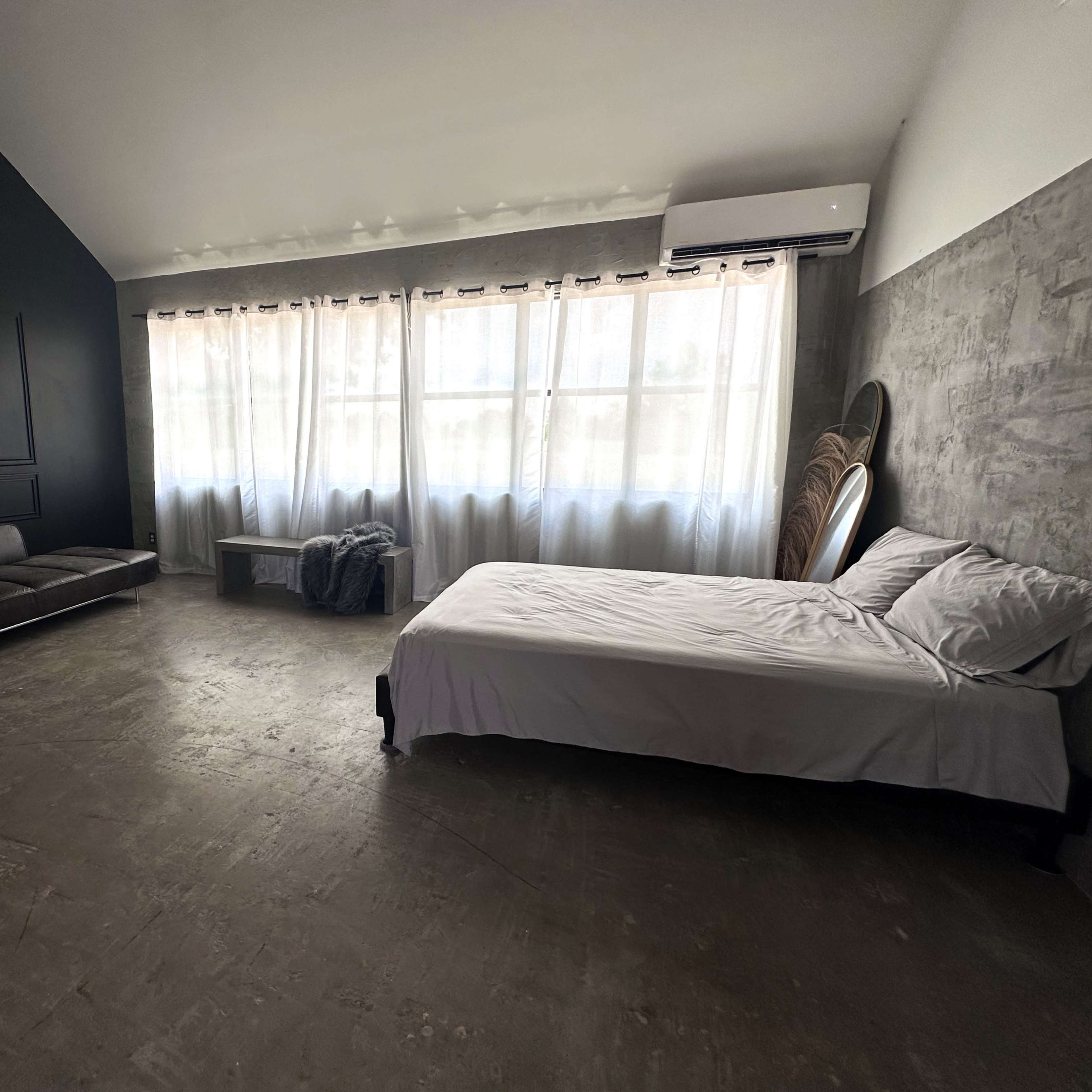 A modern bedroom features a bed, a sleek sofa, a coffee table, and large windows covered with sheer curtains, all set against dark and light walls.
