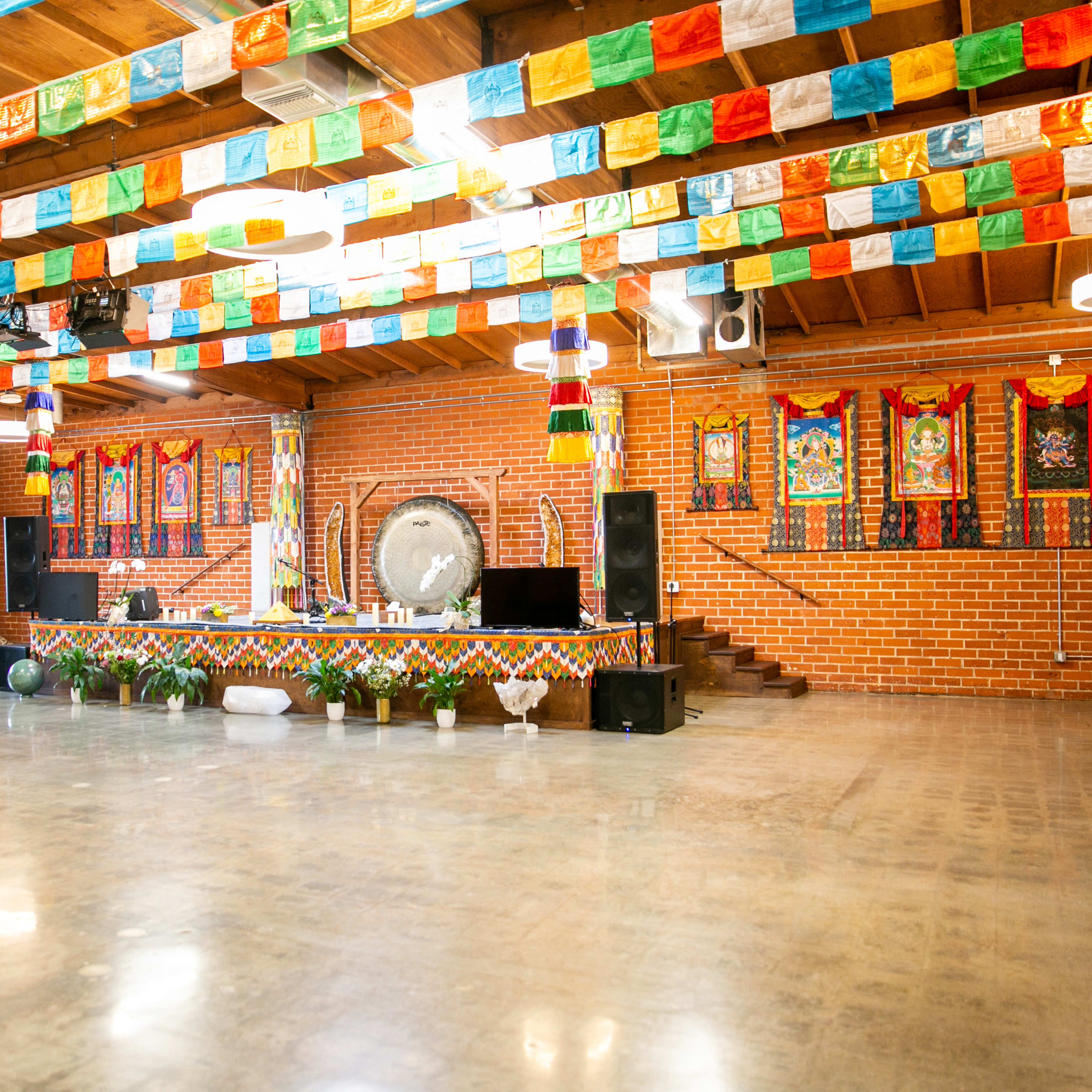 A spacious indoor venue with a stage decorated with colorful banners, lush plants, and a large gong, all set against brick walls and wooden beams.