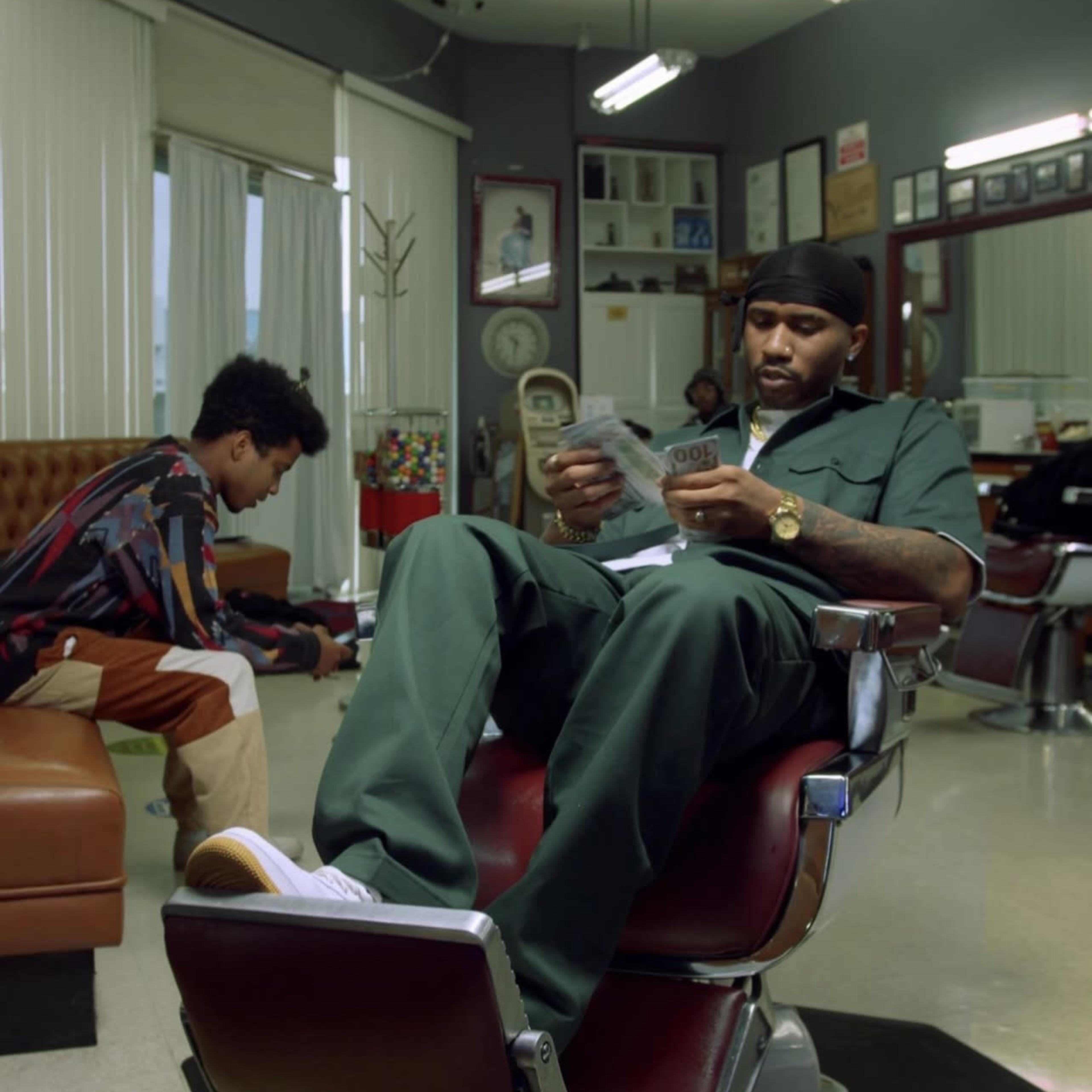 A man sits in a barber chair reading a magazine while a boy plays on the floor in a barbershop.