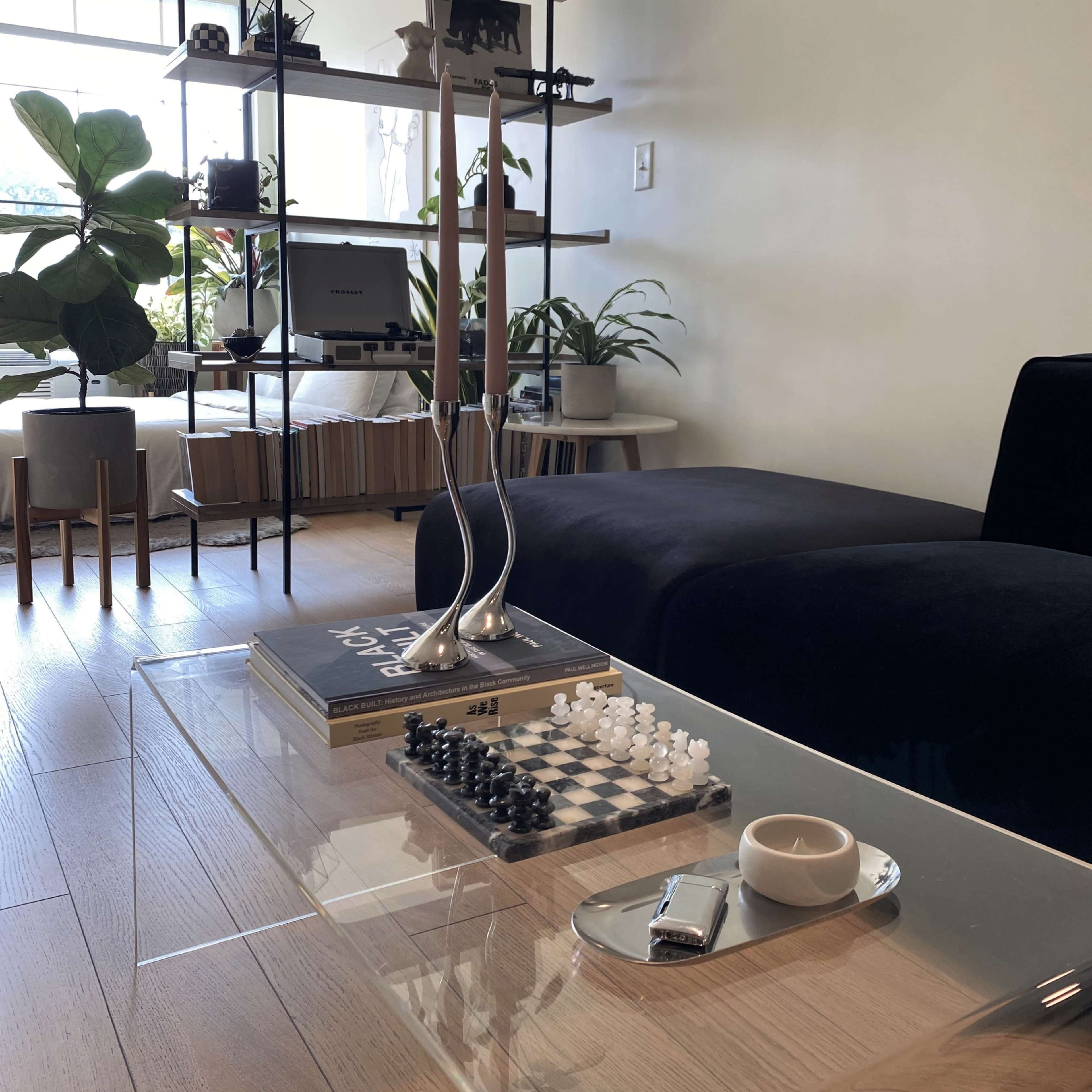 A modern living room features a glass coffee table with a chess set, a book, and a plate, surrounded by plants and stylish furniture.