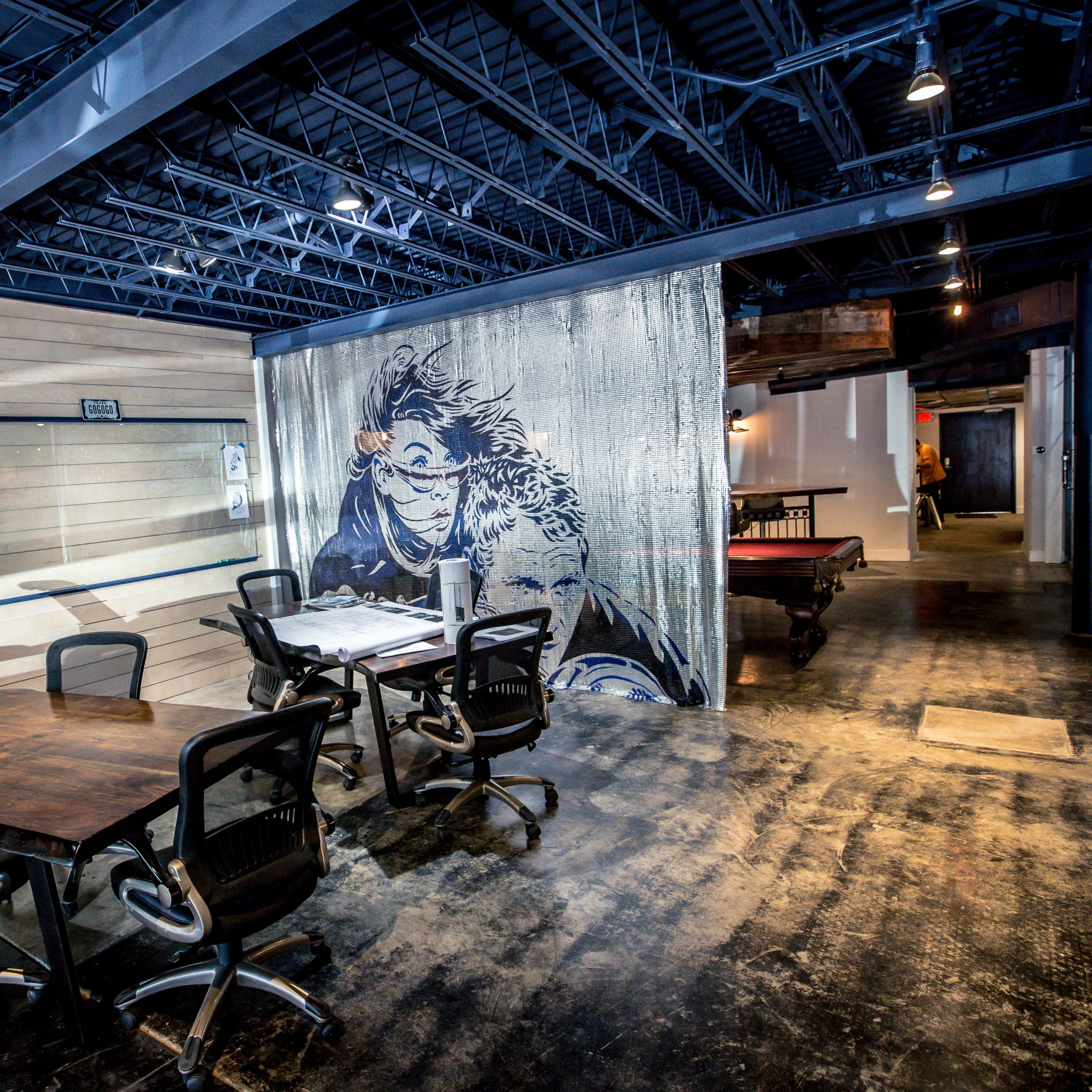 A modern office space featuring a meeting area with wooden tables and black chairs, alongside a mural and a billiard table.