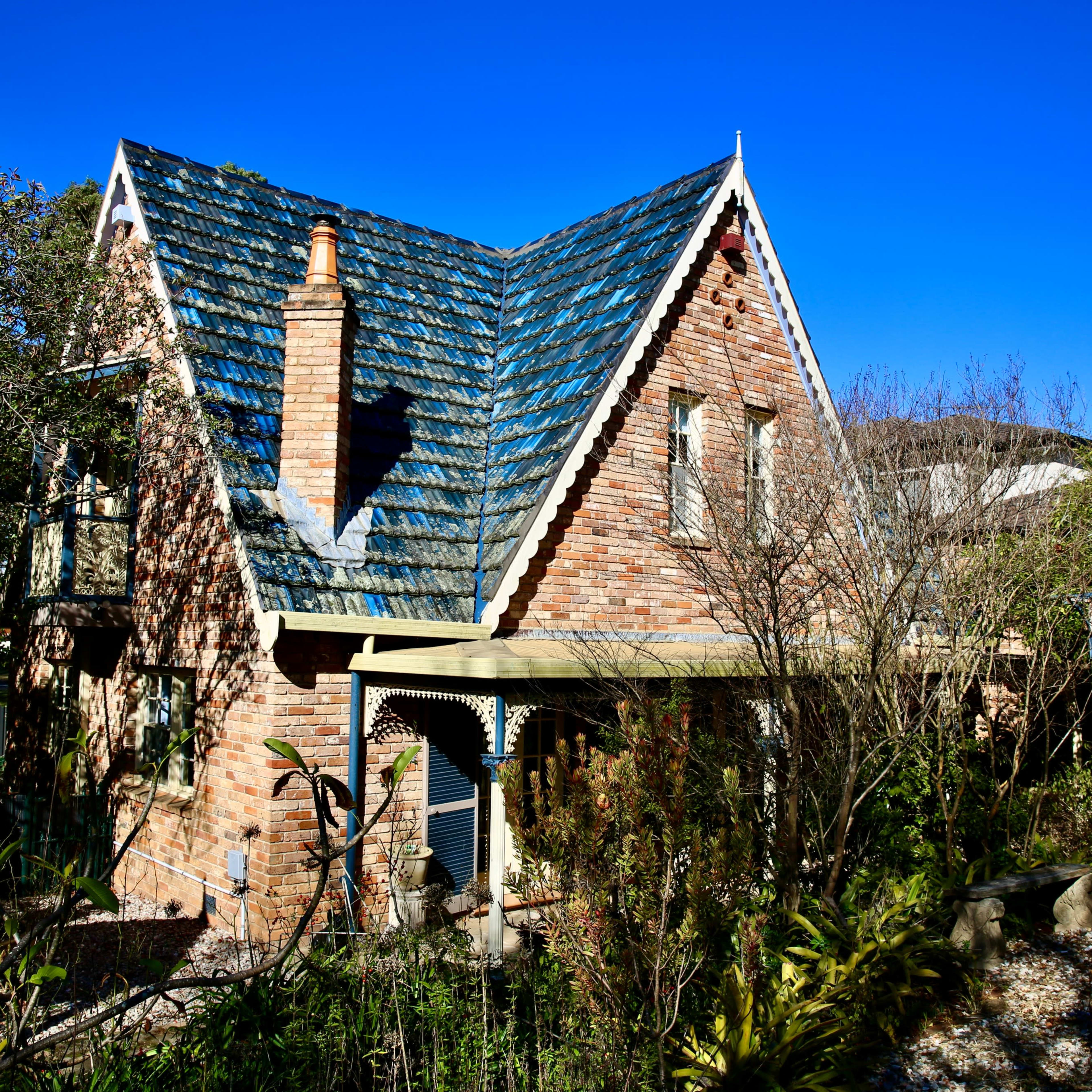 A brick house with a triangular roof, surrounded by trees and plants, is set against a clear blue sky.