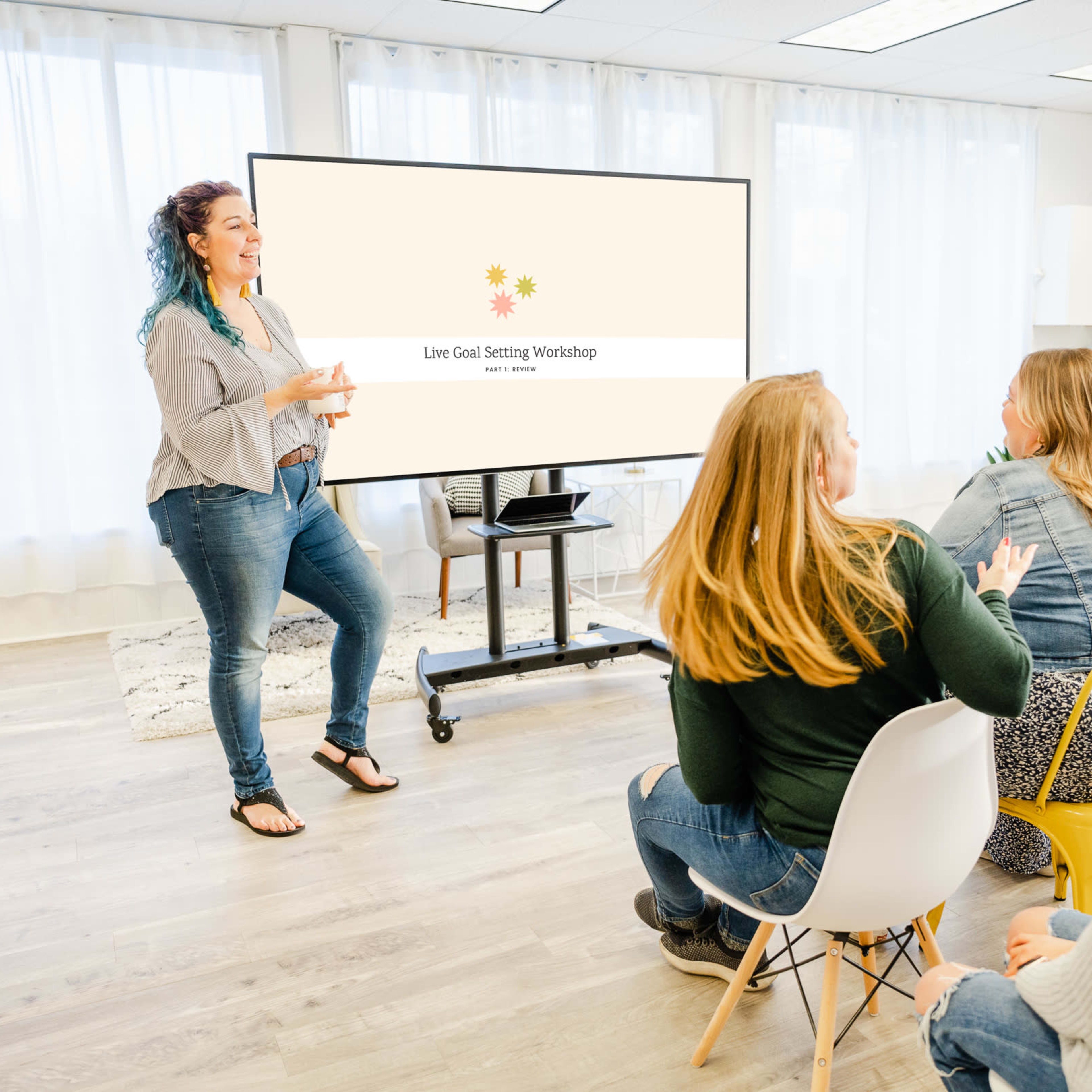 A woman with blue hair leads a goal-setting workshop in front of an audience seated on colorful chairs.
