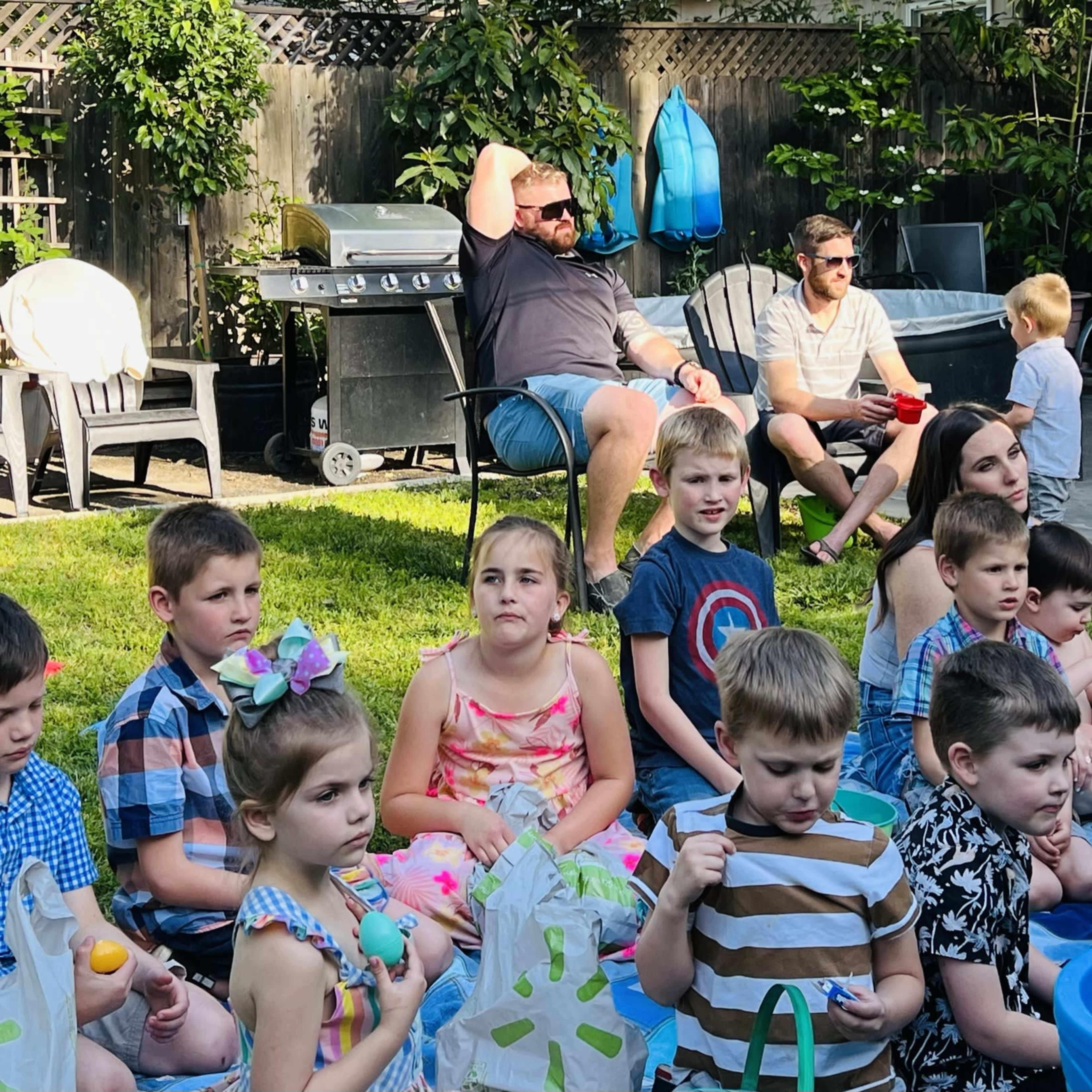 A group of children sits on the grass while adults converse in the background at a backyard gathering.