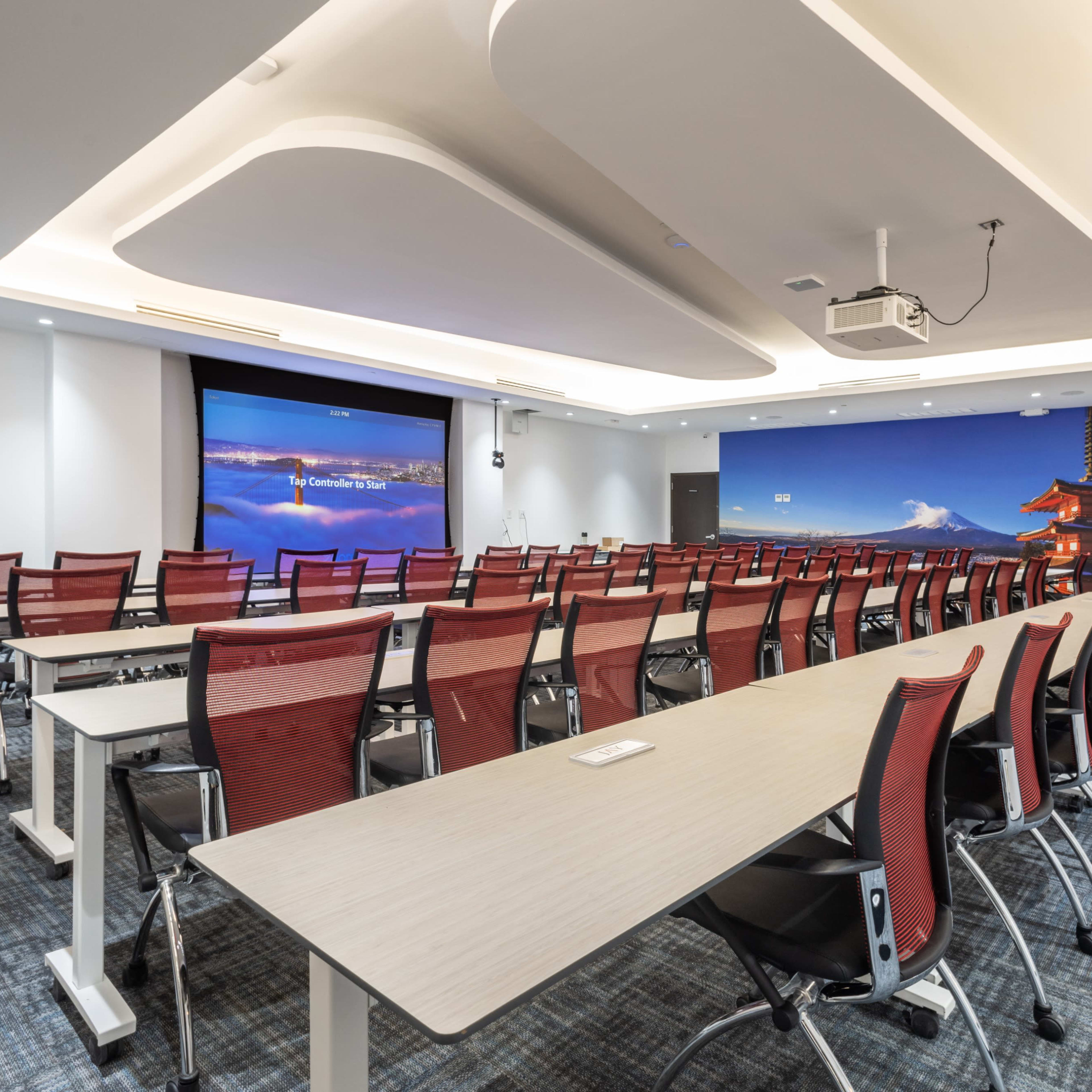 A modern conference room with rows of red chairs and long tables, featuring a large screen at one end and a scenic mural on the wall.