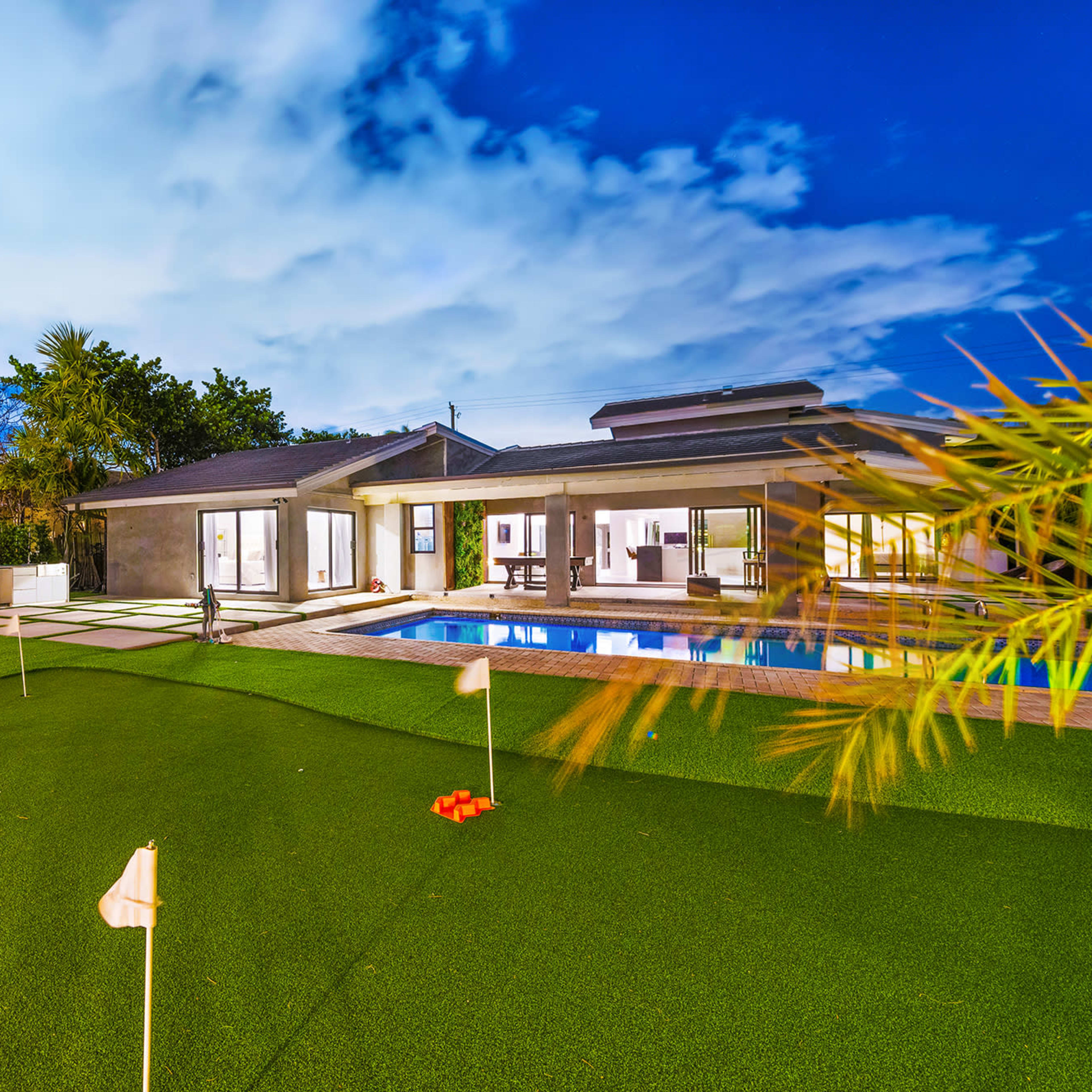 A modern house with a swimming pool and a green, manicured lawn featuring putting greens and landscaping under a blue sky.