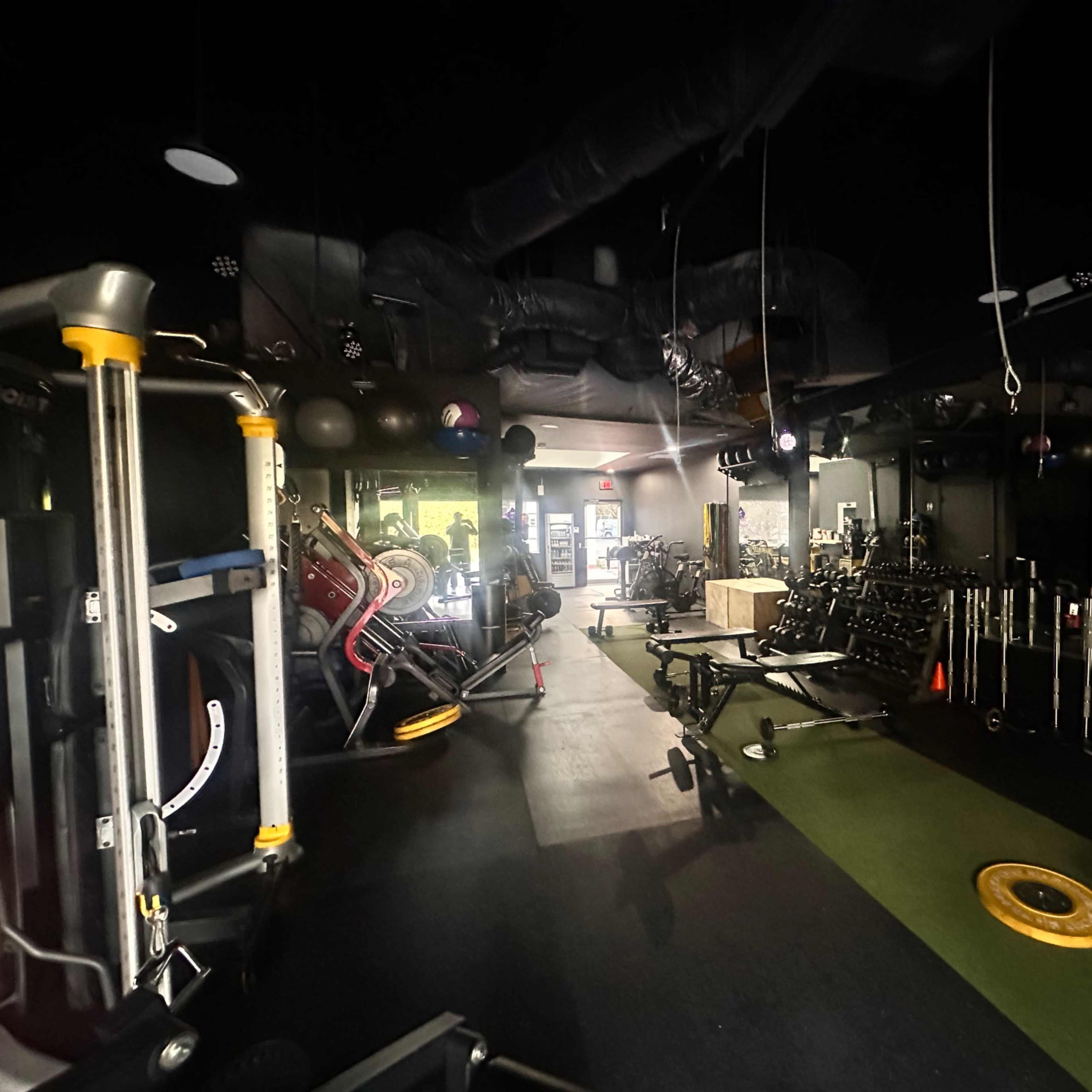 A dimly lit gym with various exercise equipment arranged along the walls and a green turf area in the center.