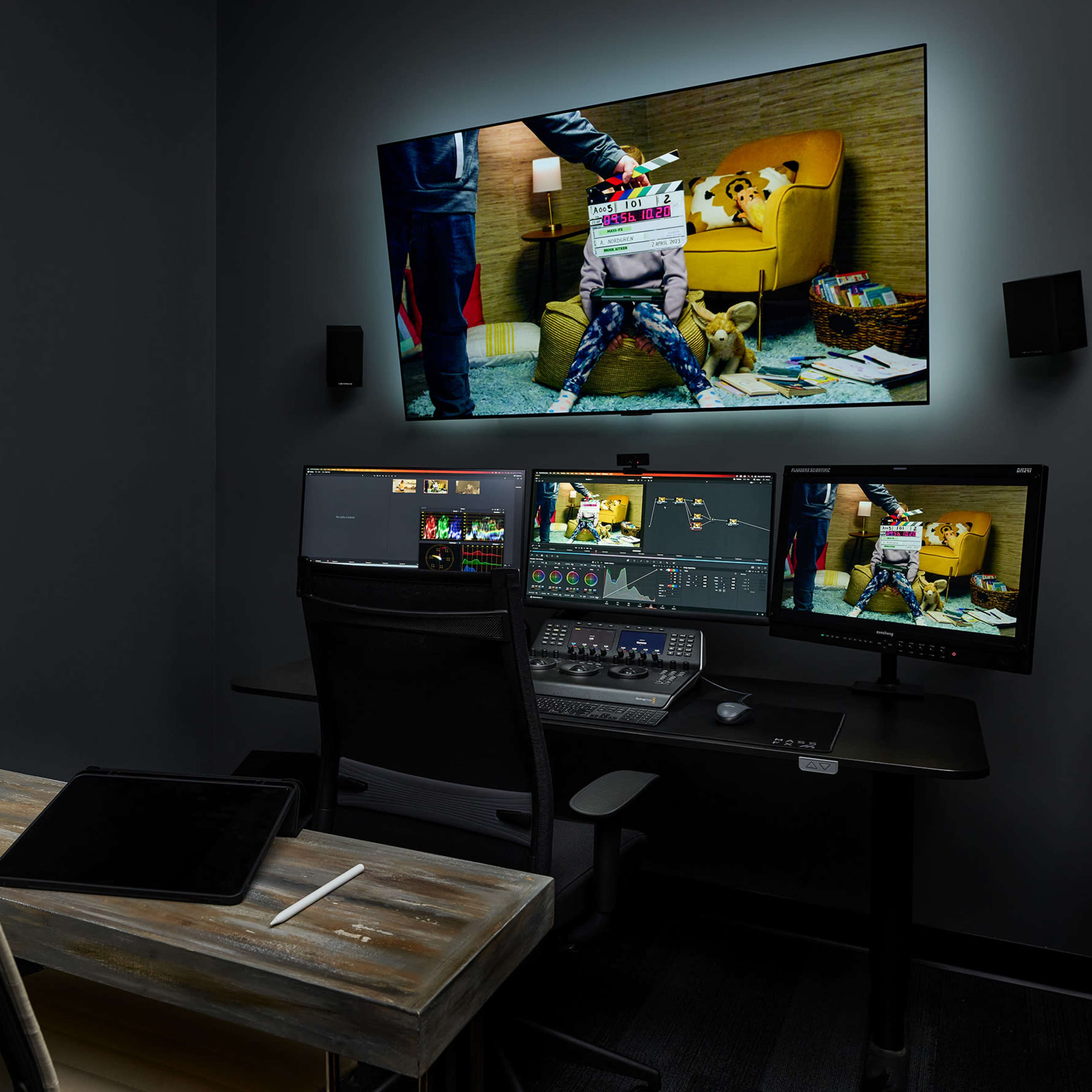 A video editing suite features multiple monitors, a control panel, and a wall-mounted screen displaying a scene with a child holding a clapperboard.