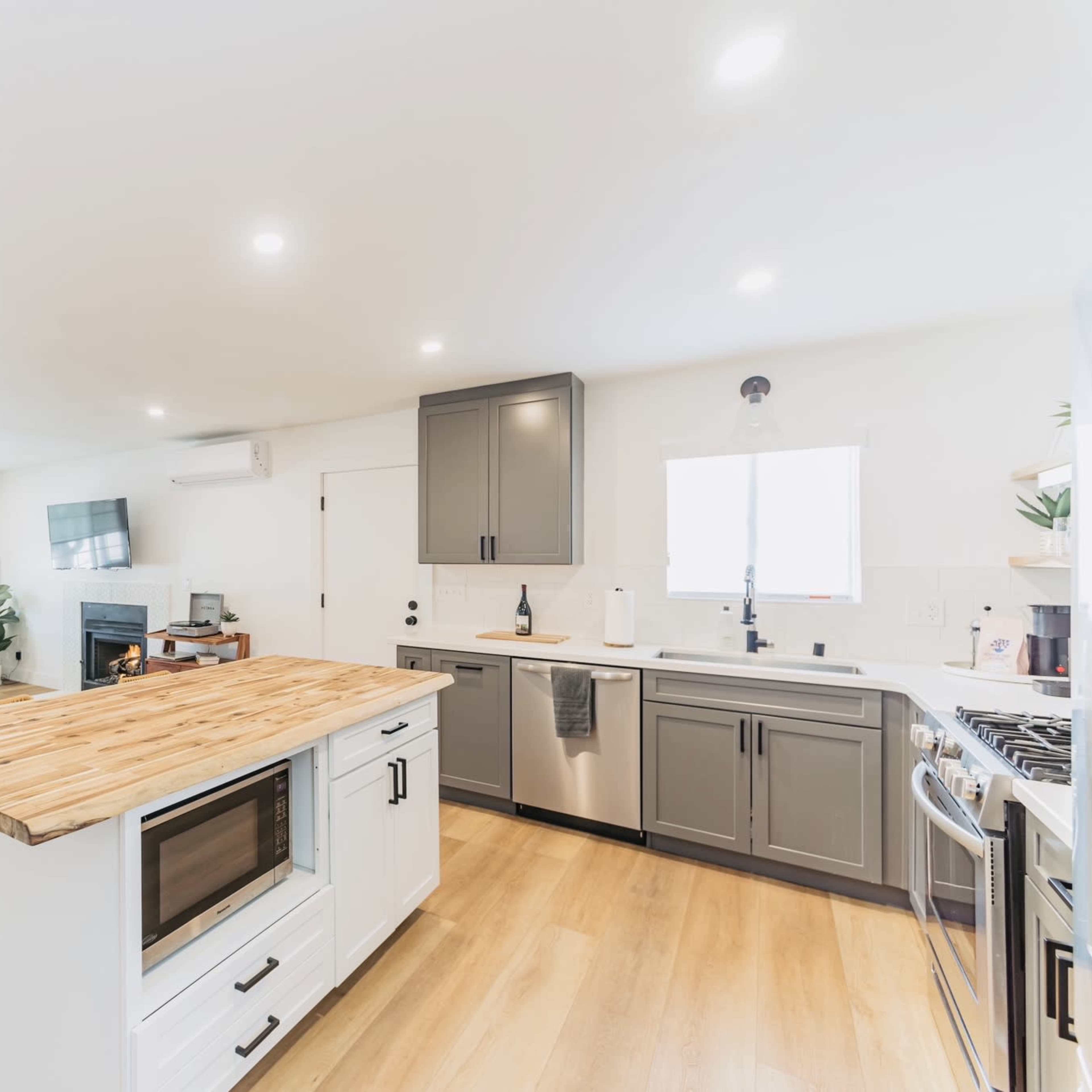 A modern kitchen and living space featuring an island with a wooden countertop, grey cabinetry, and a comfortable seating area with a light-colored couch and a brown chair.