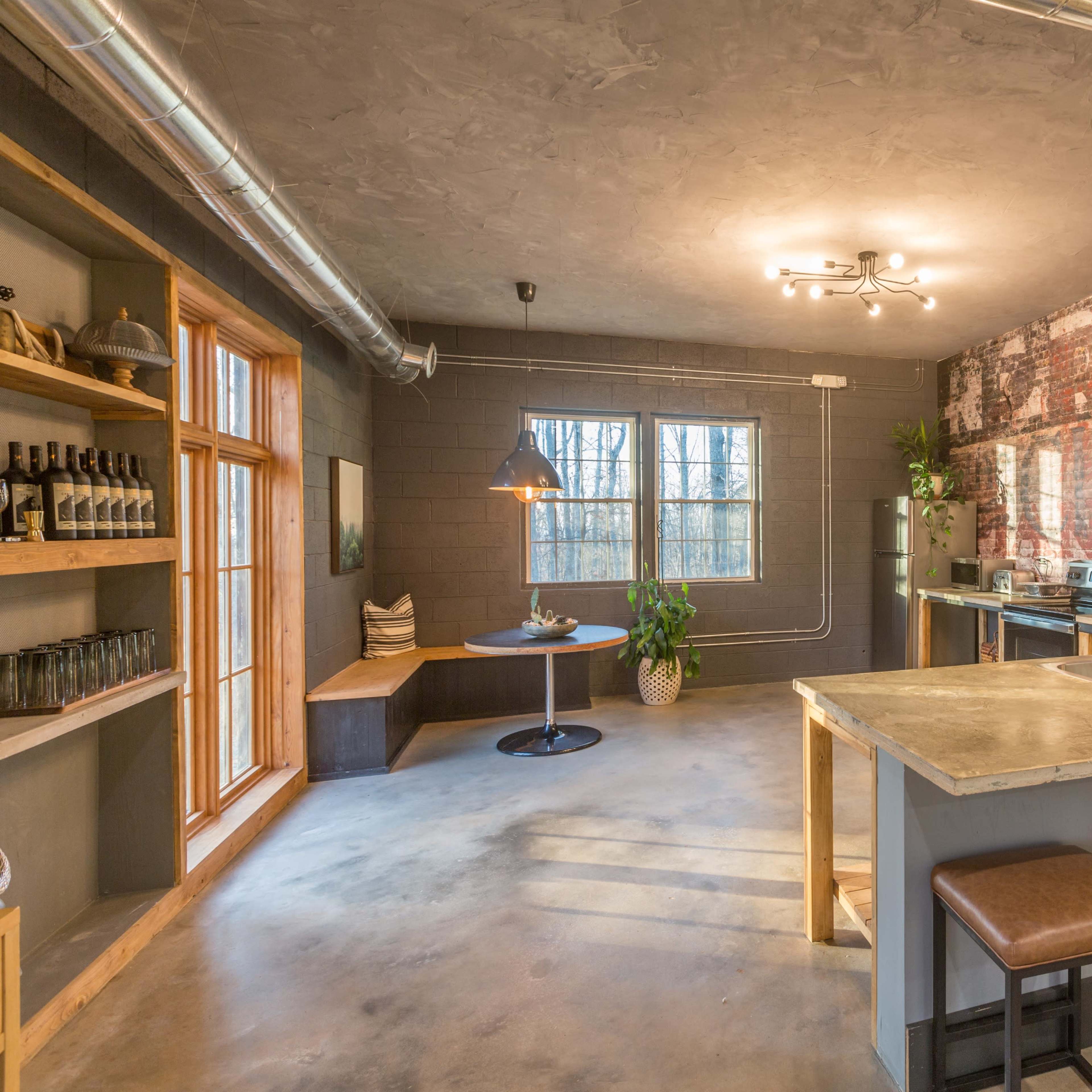 A modern kitchen with a concrete countertop, bar seating, and exposed brick walls, featuring large windows that overlook a wooded area.
