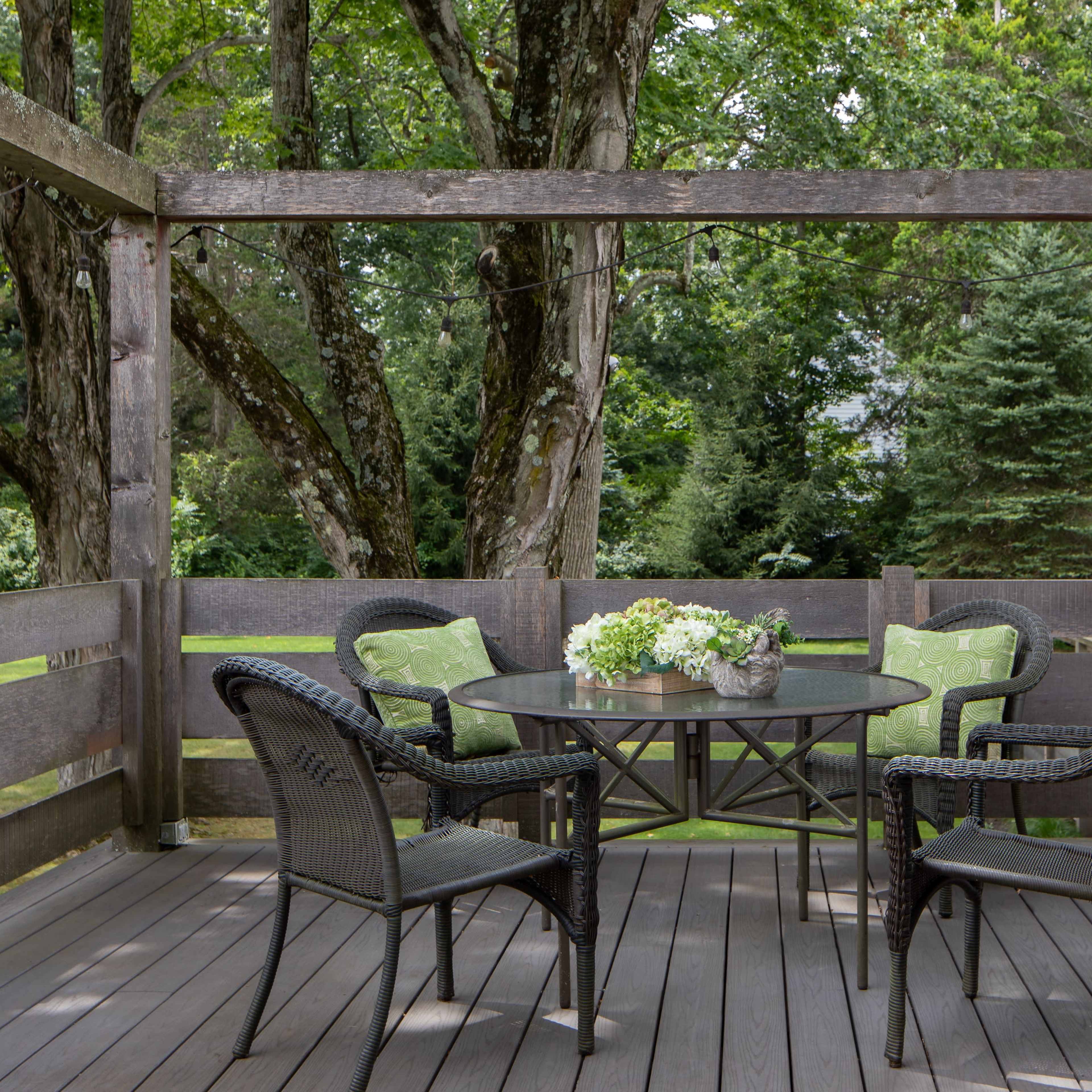 A wooden deck features a round table surrounded by four wicker chairs, with green cushions and potted plants on the table.