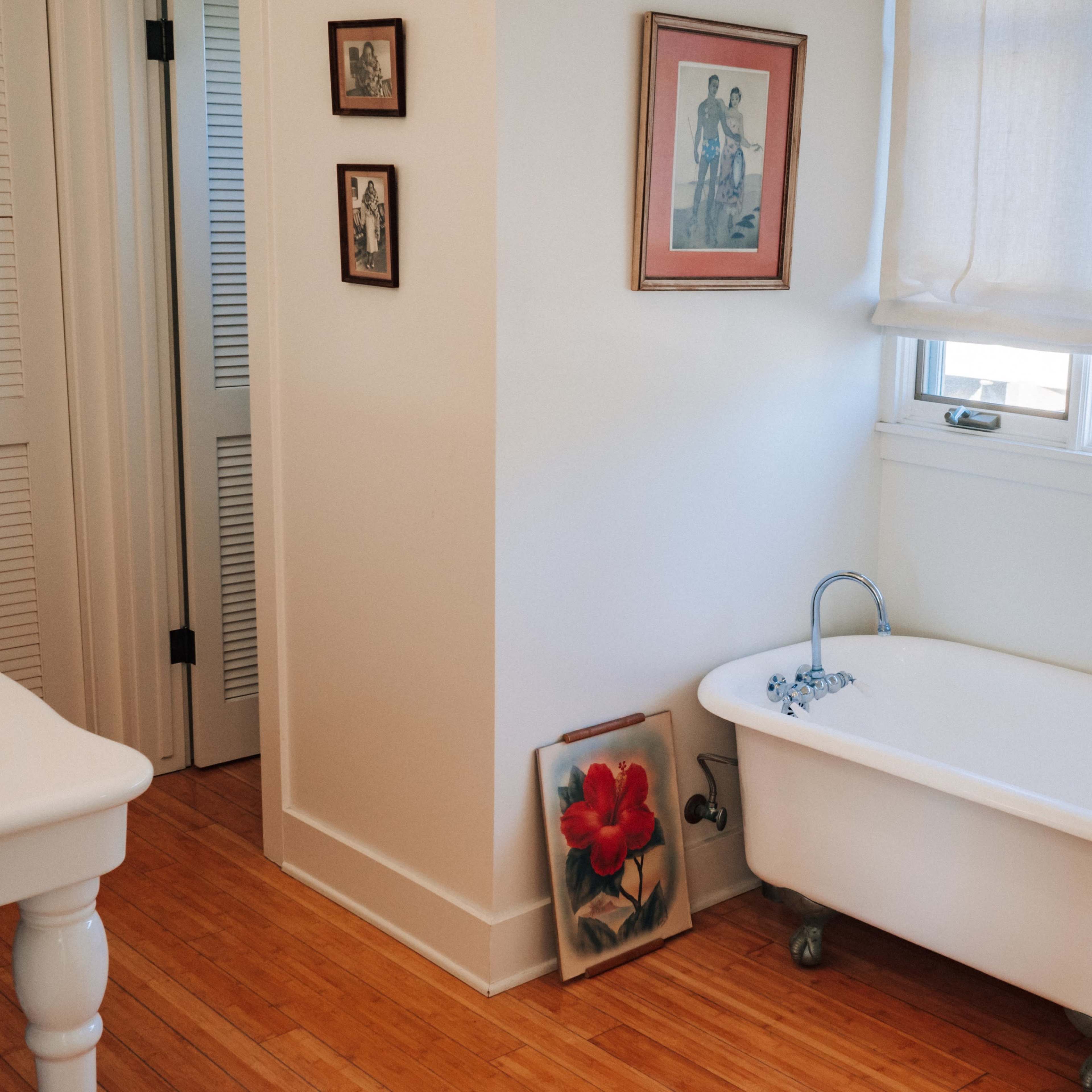 A bright bathroom featuring a freestanding bathtub, a framed floral painting on the floor, and framed pictures on the wall.