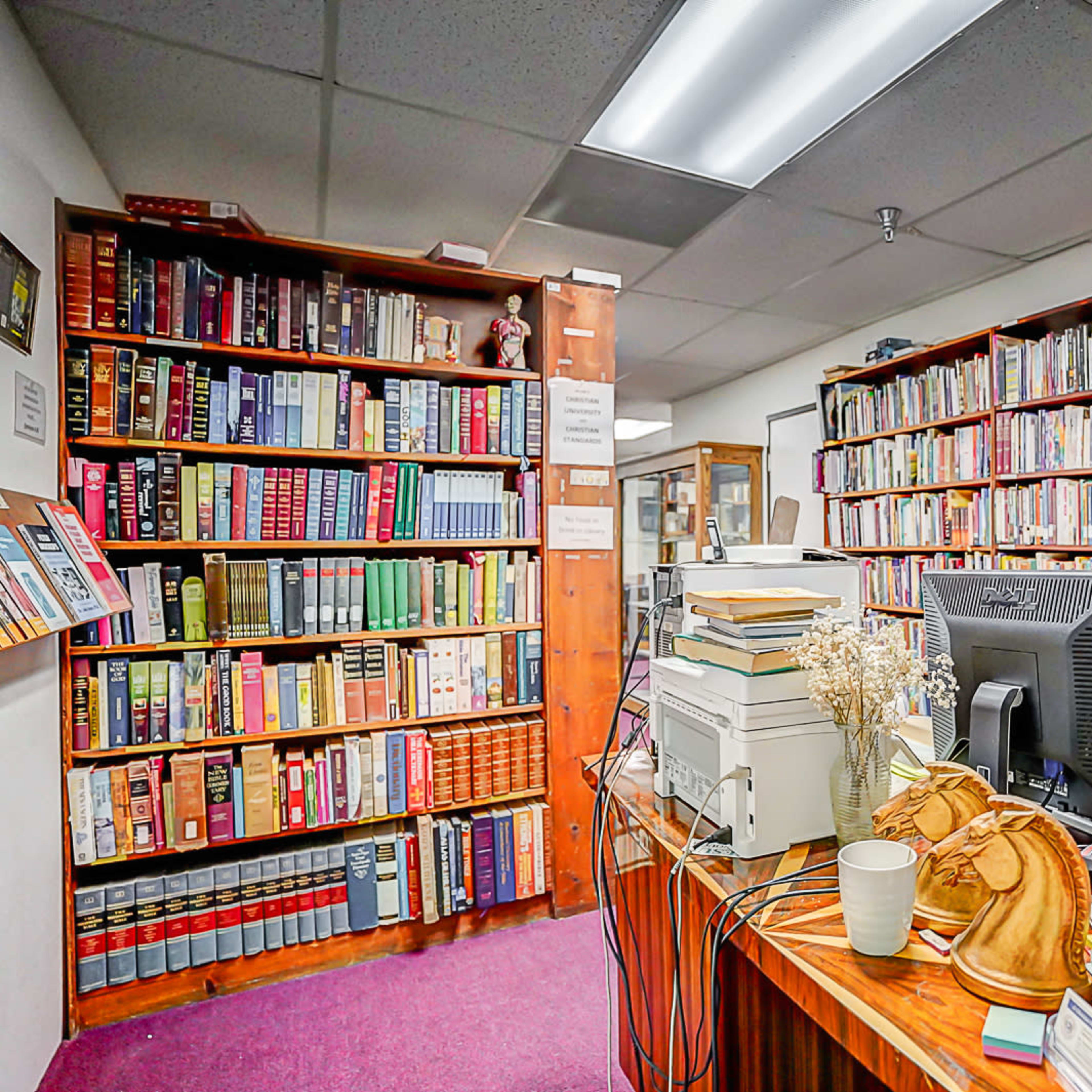 The scene features a study or office filled with bookshelves lined with various books, a desk cluttered with office supplies, and a computer monitor on the right side.