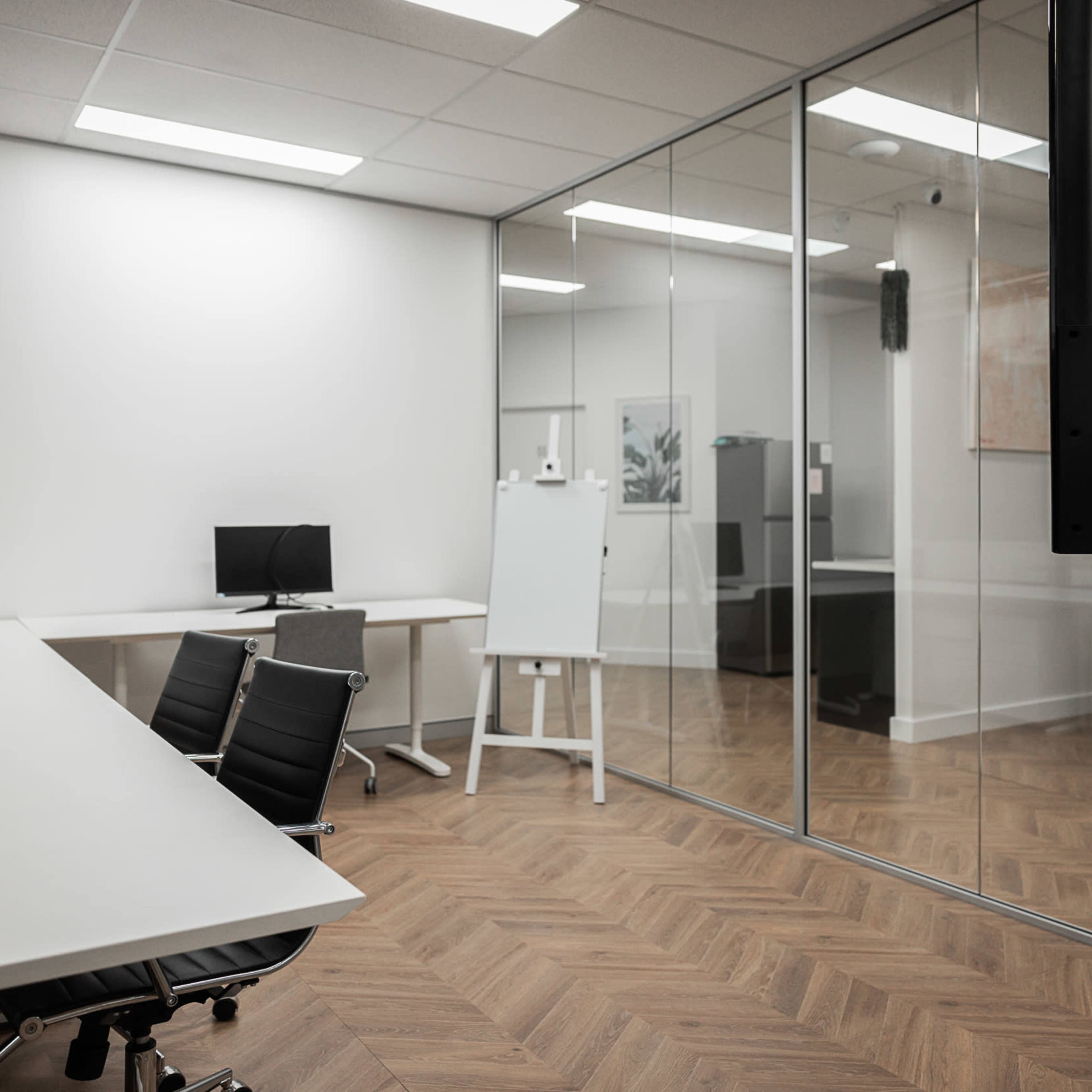 A modern office space with a glass-walled meeting room, two workstations with computers, and a whiteboard.