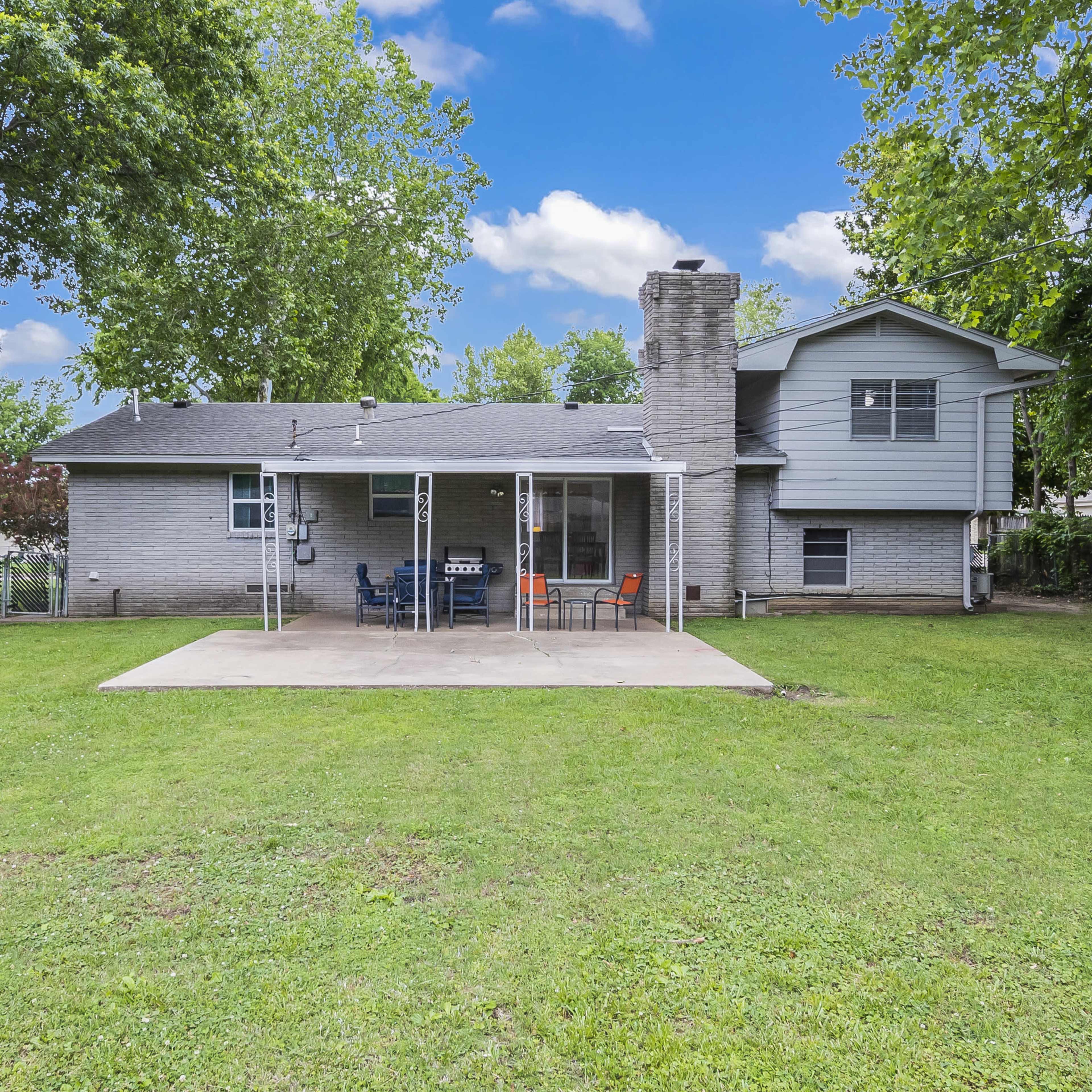 A two-story house with a brick exterior, a chimney, and a concrete patio in the backyard, surrounded by grass and trees.