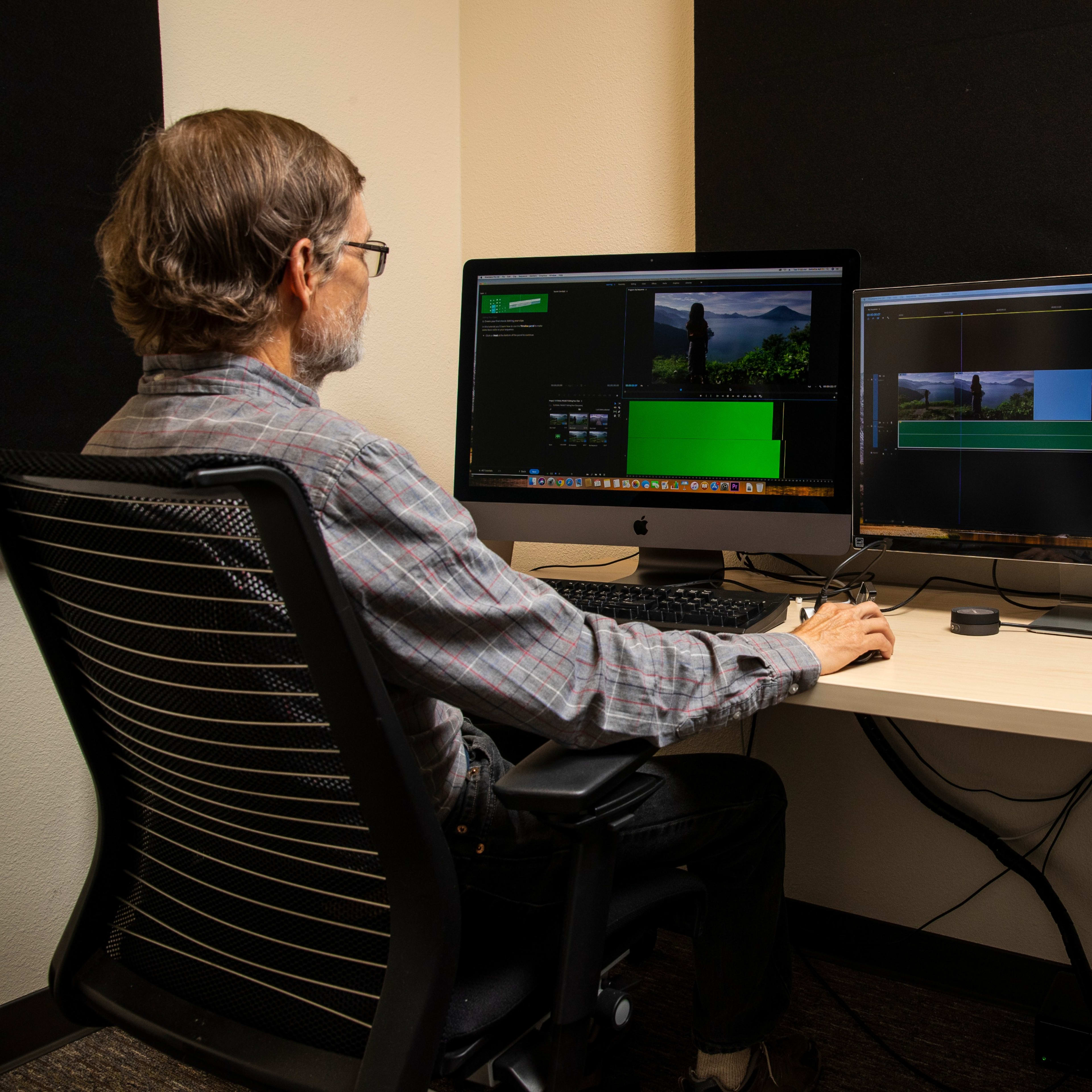 A man sits at a desk in front of two computer monitors editing video, with a sign to the right providing instructions for using the edit suite.