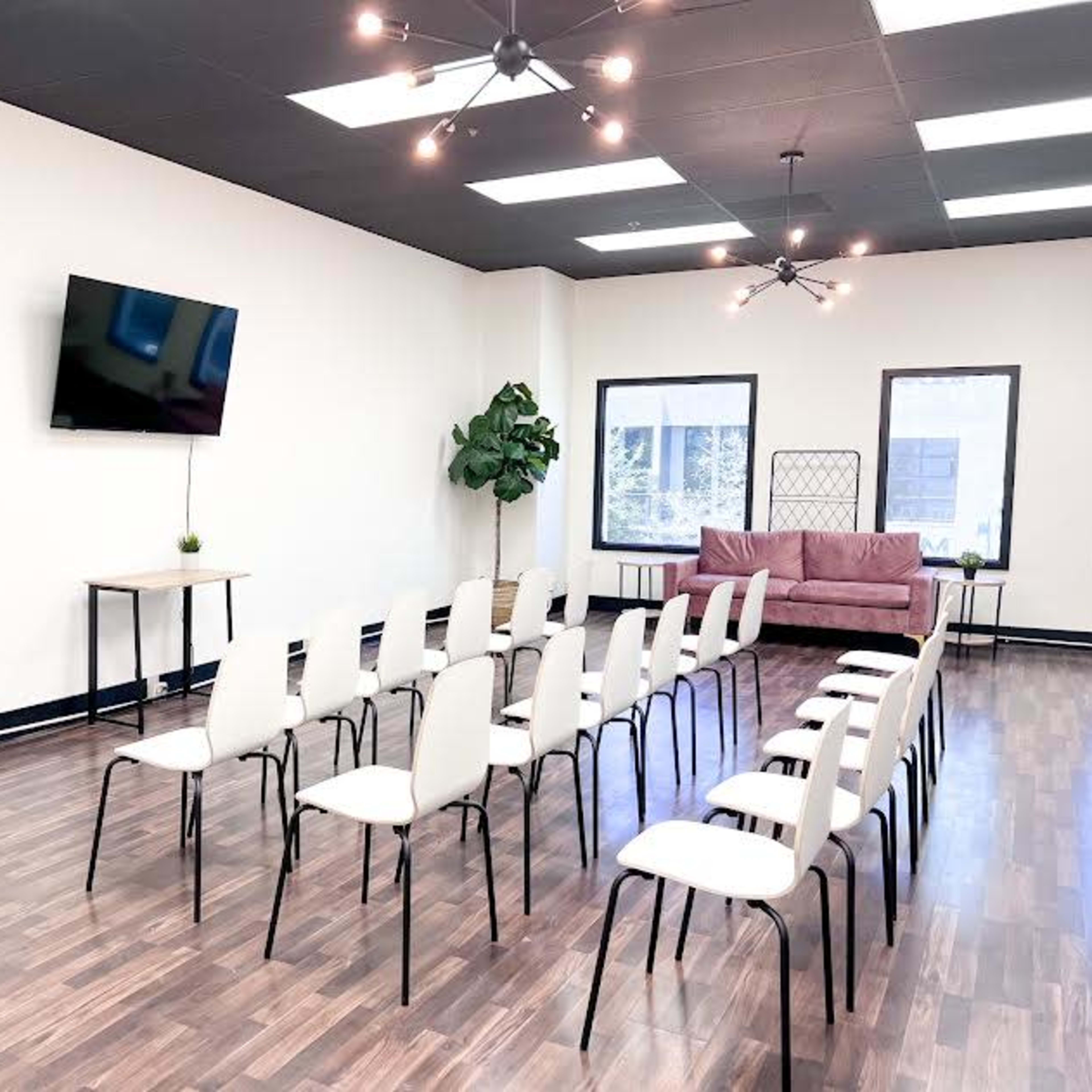 A bright meeting room is set up with a row of white chairs facing a large television and a whiteboard, complemented by a pink sofa and greenery.