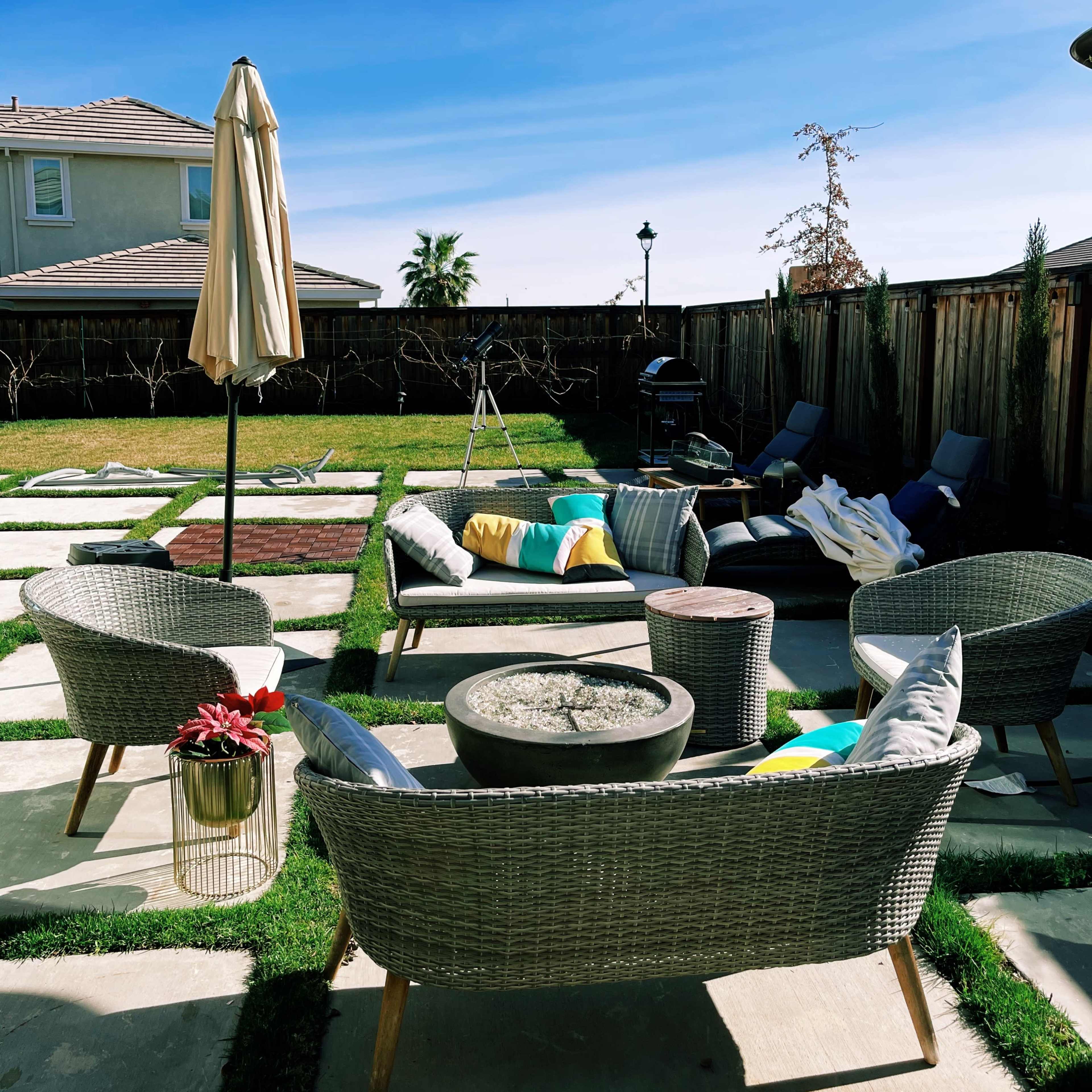 A backyard patio features wicker seating arranged around a fire pit, with decorative pillows and a sun umbrella beside a grassy area and pavers leading to a wooden deck.
