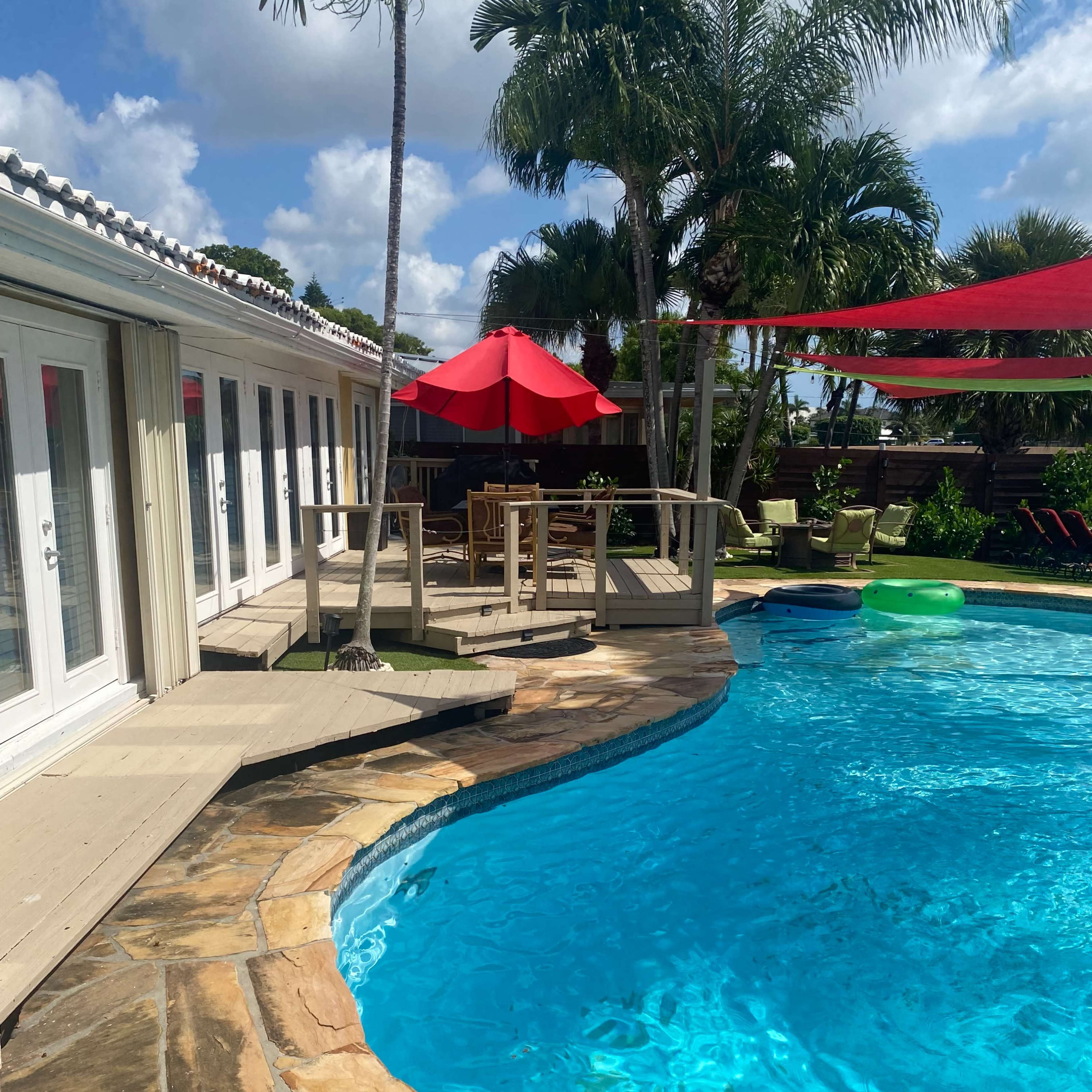 A swimming pool surrounded by a wooden deck, lounge chairs, and a shaded red umbrella with palm trees in the background.